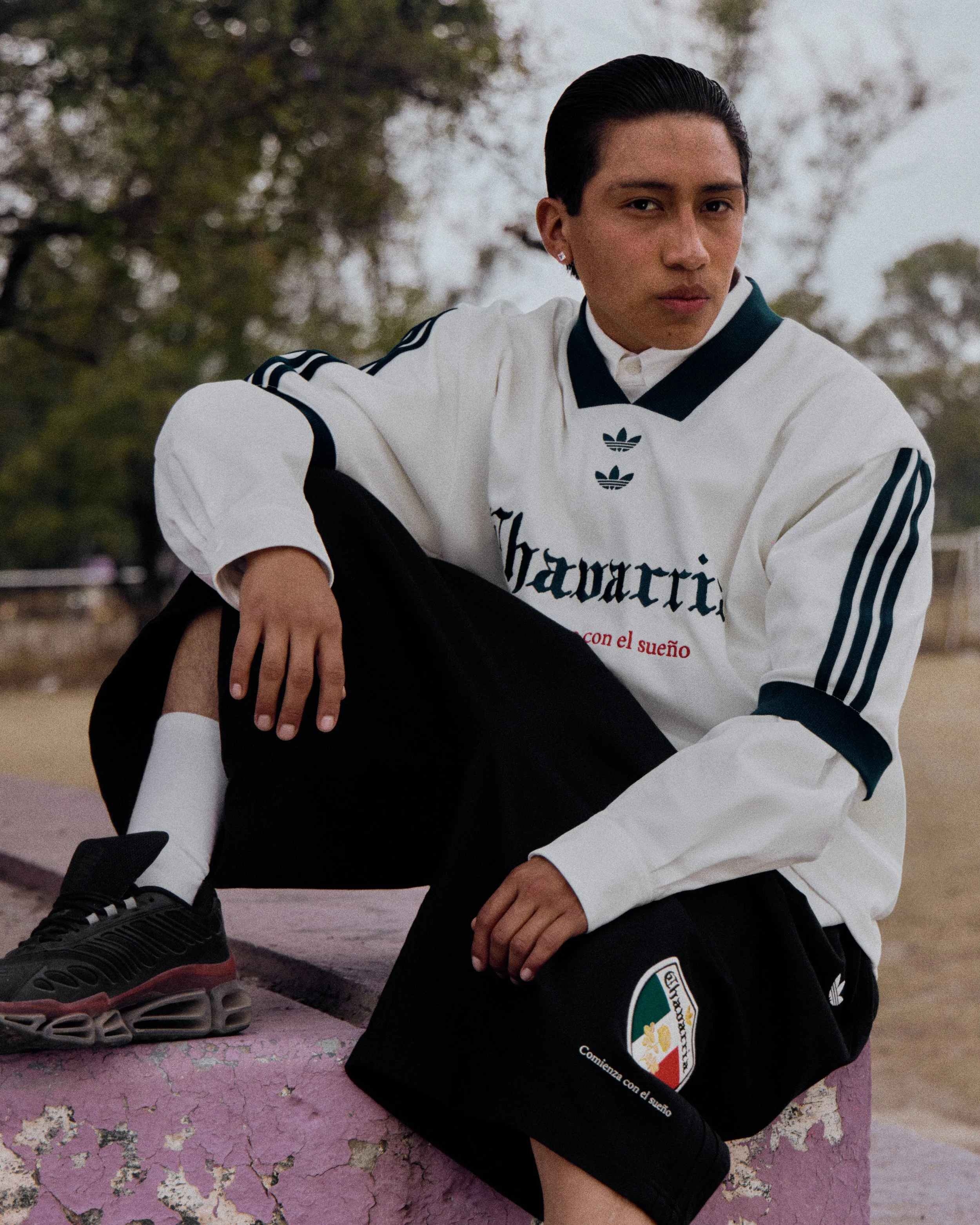 A young man with short dark hair wearing a white and black Adidas sports jacket, black shorts with patches, and white socks, sitting on a cracked pink surface outdoors with trees in the background.