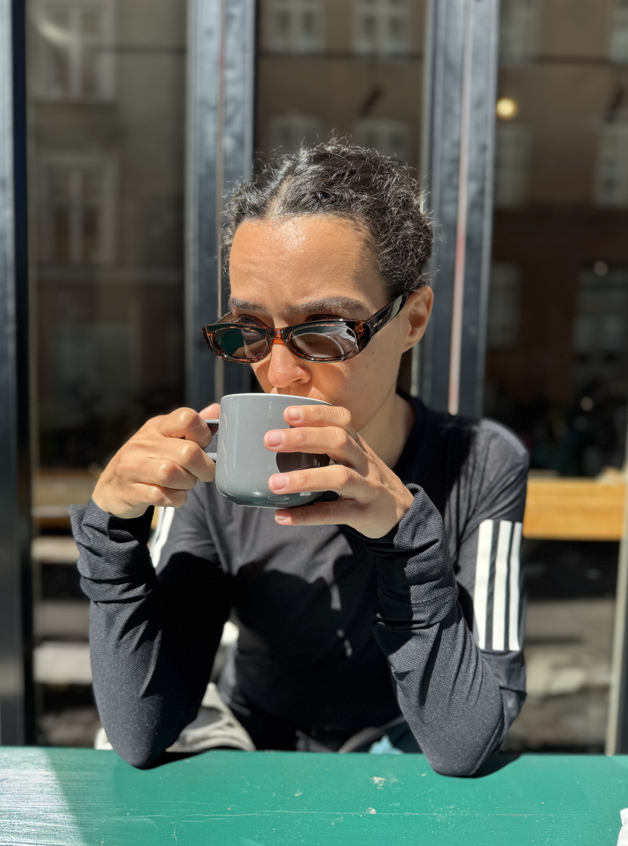 Woman with dark curly hair wearing sunglasses and a black athletic jacket, drinking from a gray mug outdoors at a sunny cafe or patio.