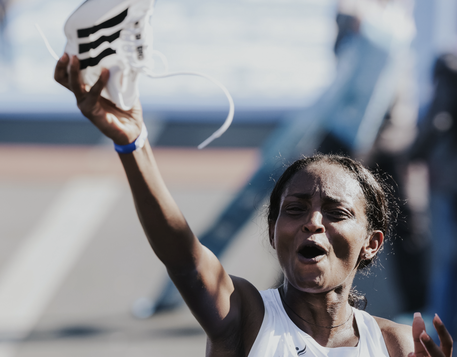 Young woman with wet hair raising her arm, holding a crumpled white and black Adidas shoe during an outdoor sports event.