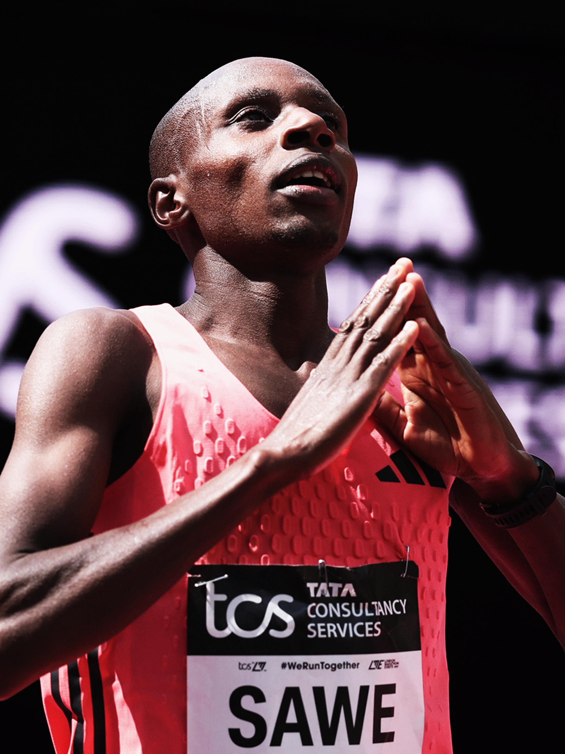 A female marathon runner with a shaved head, wearing a pink athletic top, praying with her hands pressed together in front of her chest during a race.