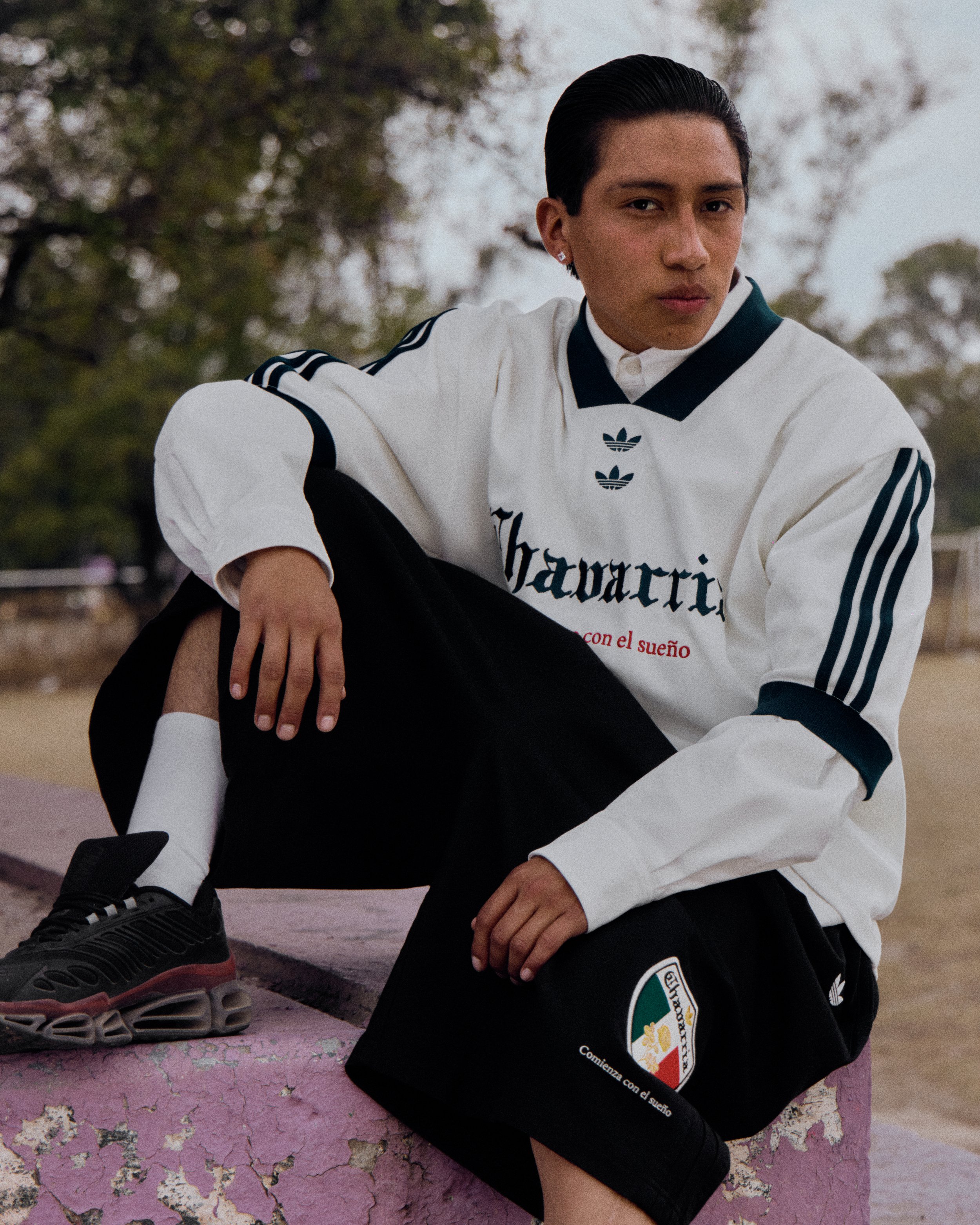 A young person with short dark hair, wearing a white sports jacket with black and green accents, seated outdoors on a pink, weathered surface, with trees in the background.