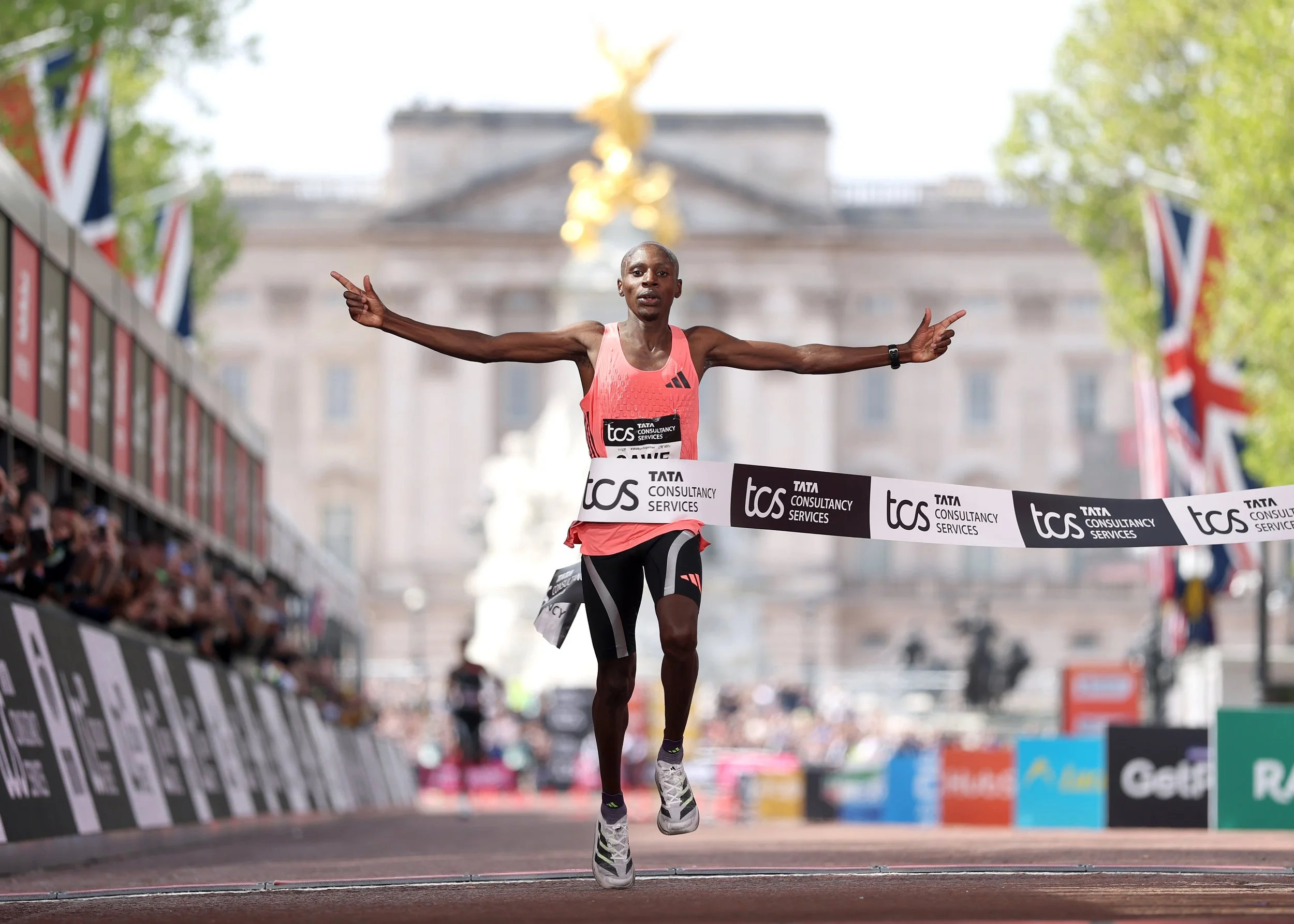 A male marathon runner crossing the finish line with his arms outstretched, wearing a pink tank top, black shorts, and white running shoes, with his head bald, surrounded by a crowd and flags, and a historic building in the background.