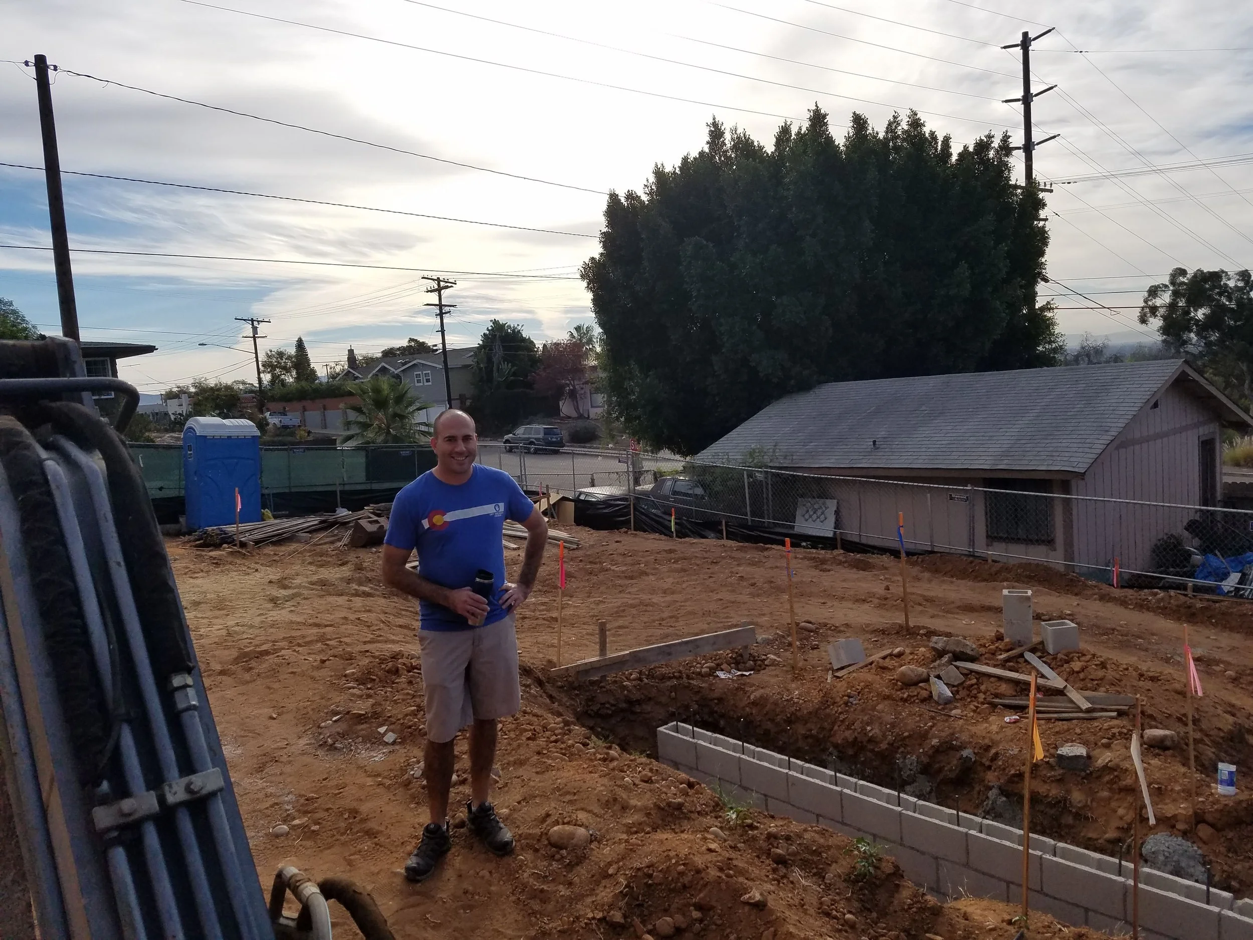 A man wearing a blue T-shirt and khaki shorts stands at a construction site with dirt, wooden planks, and concrete blocks, smiling with a drink in his hand. In the background are houses, utility poles, trees, and a partly cloudy sky.