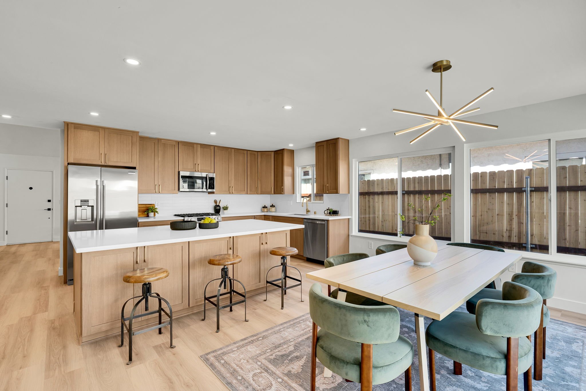 Modern kitchen with wooden cabinets, stainless steel appliances, a kitchen island with stools, a dining table with green velvet chairs, large windows, and a contemporary chandelier.