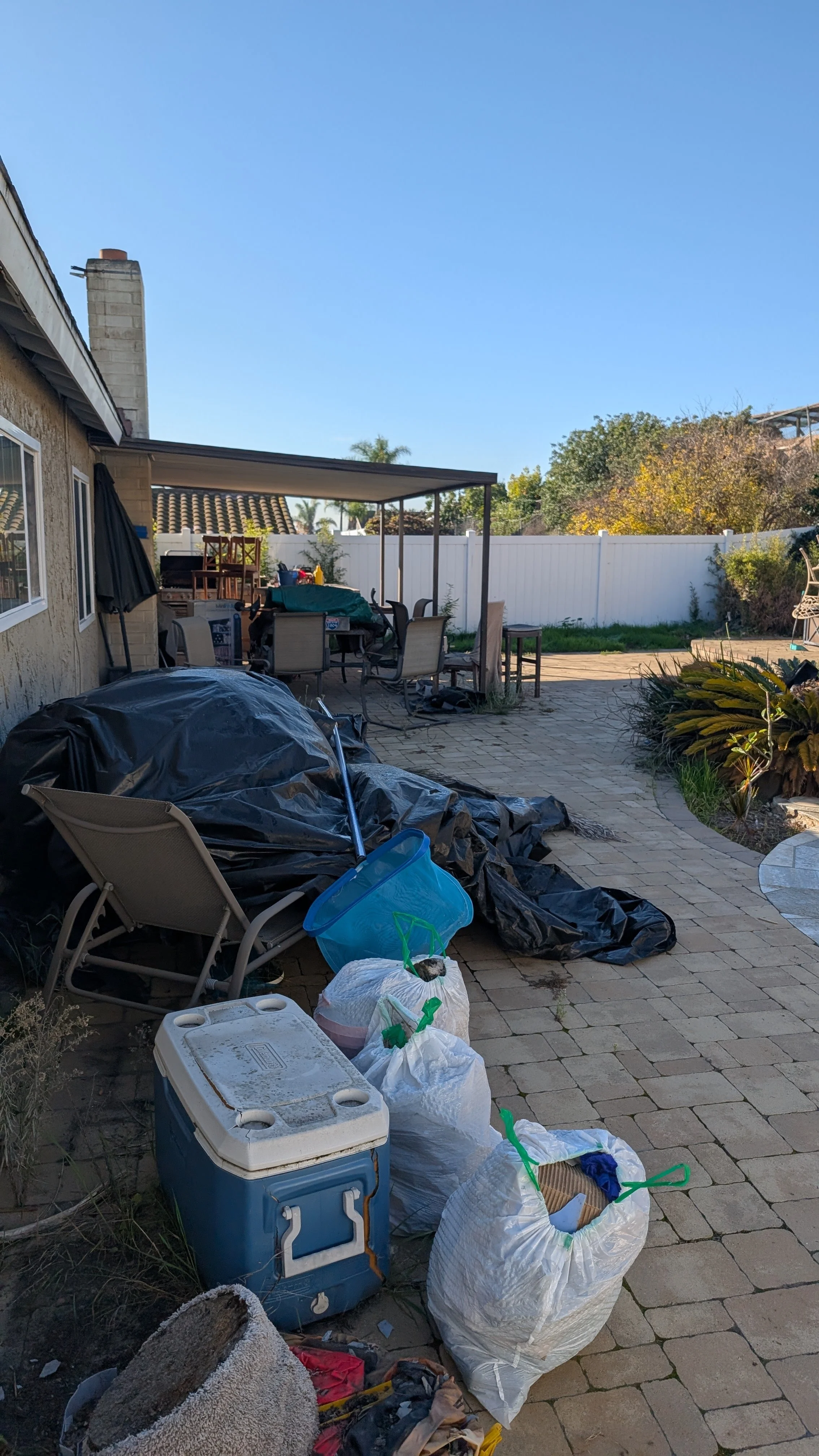 Backyard with clutter, including plastic bags, coolers, and miscellaneous items, on a brick patio under a clear blue sky.