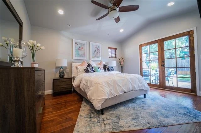 Bedroom with a large bed, nightstands with lamps, artwork above the bed, sliding glass doors leading to an outdoor patio, ceiling fan, wooden flooring, and a blue area rug.