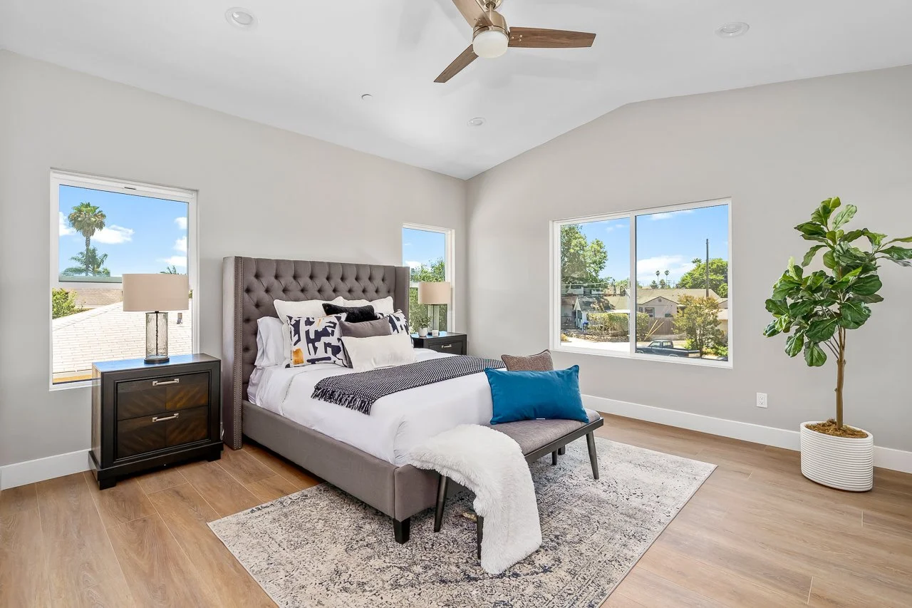 Modern bedroom with a gray tufted bed, white bedding, decorative pillows, two nightstands with lamps, a beige area rug, a large window showing a neighborhood, and a potted plant.