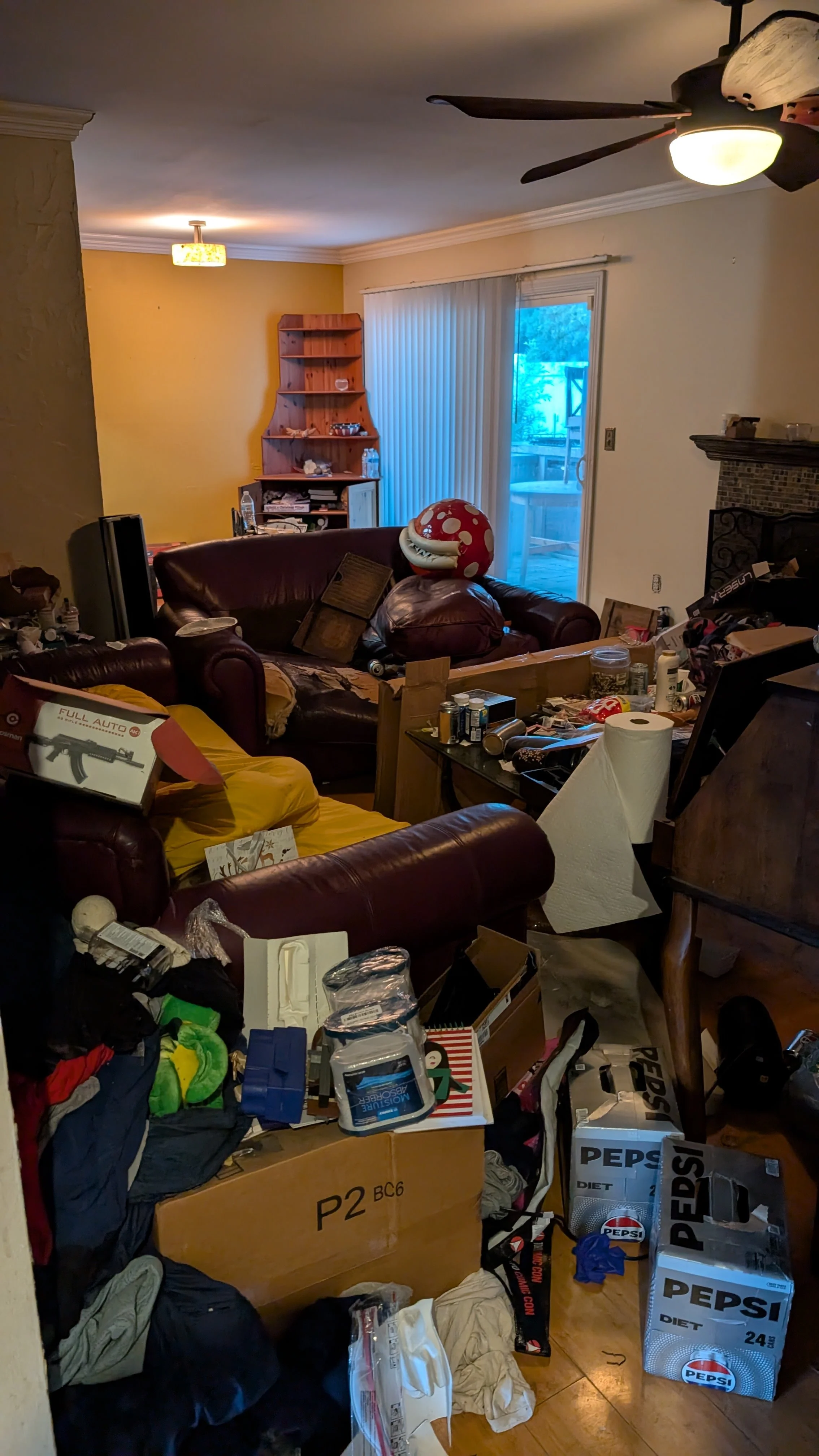 A cluttered living room filled with scattered clothes, boxes, and various household items, including a brown leather couch, a ceiling fan, and a sliding glass door with vertical blinds leading to an outdoor area.