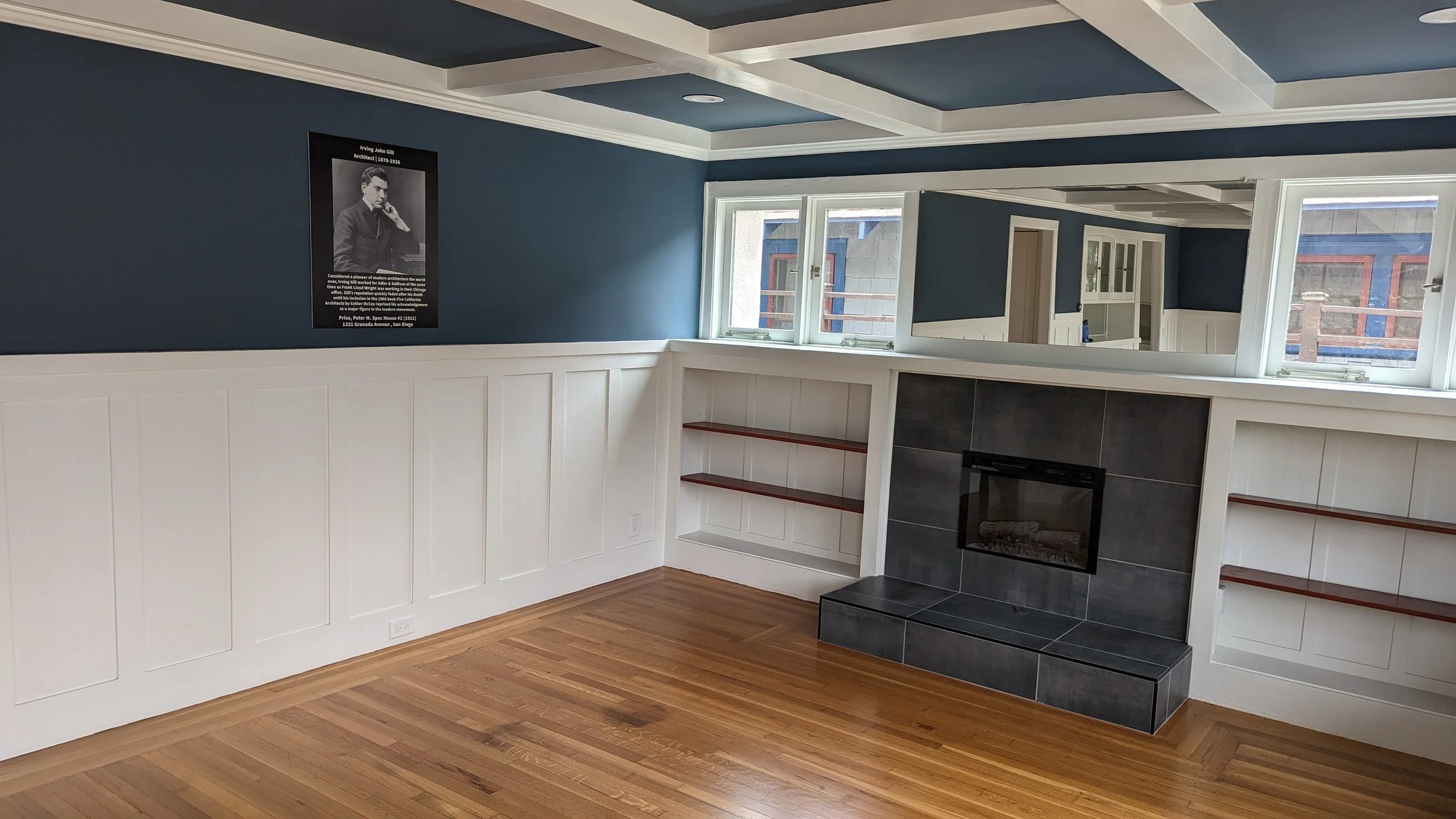 Interior view of a living room with a blue ceiling, white wainscoting, a fireplace with black tiles, and windows showing neighboring buildings.