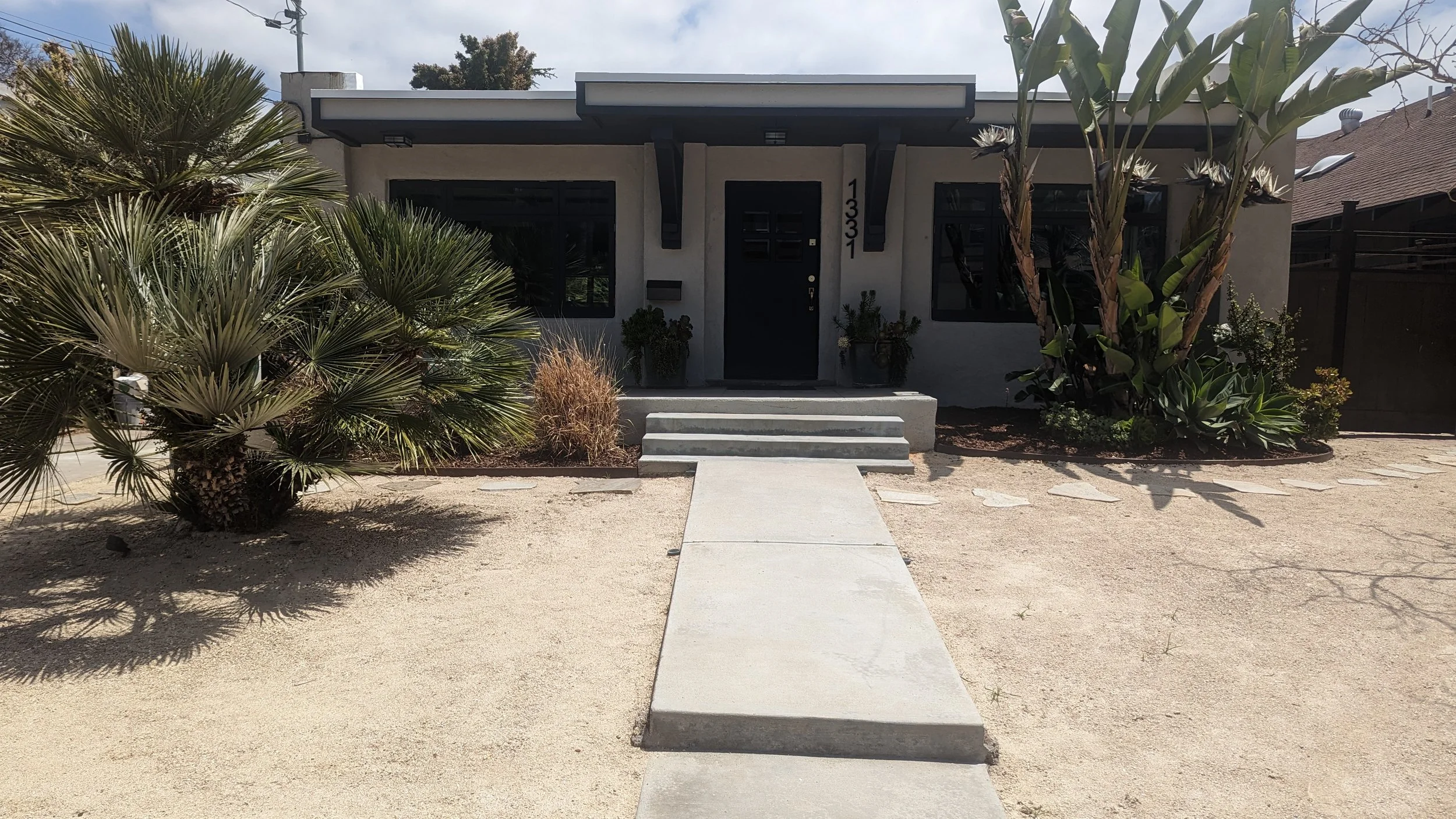 A modern single-story house with black windows and door, decorated with plants and white stairs leading to the entrance, situated in a yard with sand and minimal landscaping.