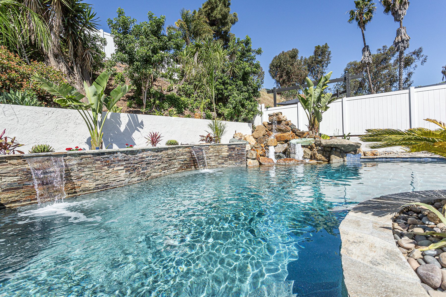 A backyard swimming pool with a stone waterfall feature, surrounded by lush green tropical plants and a white privacy fence, under a clear blue sky.