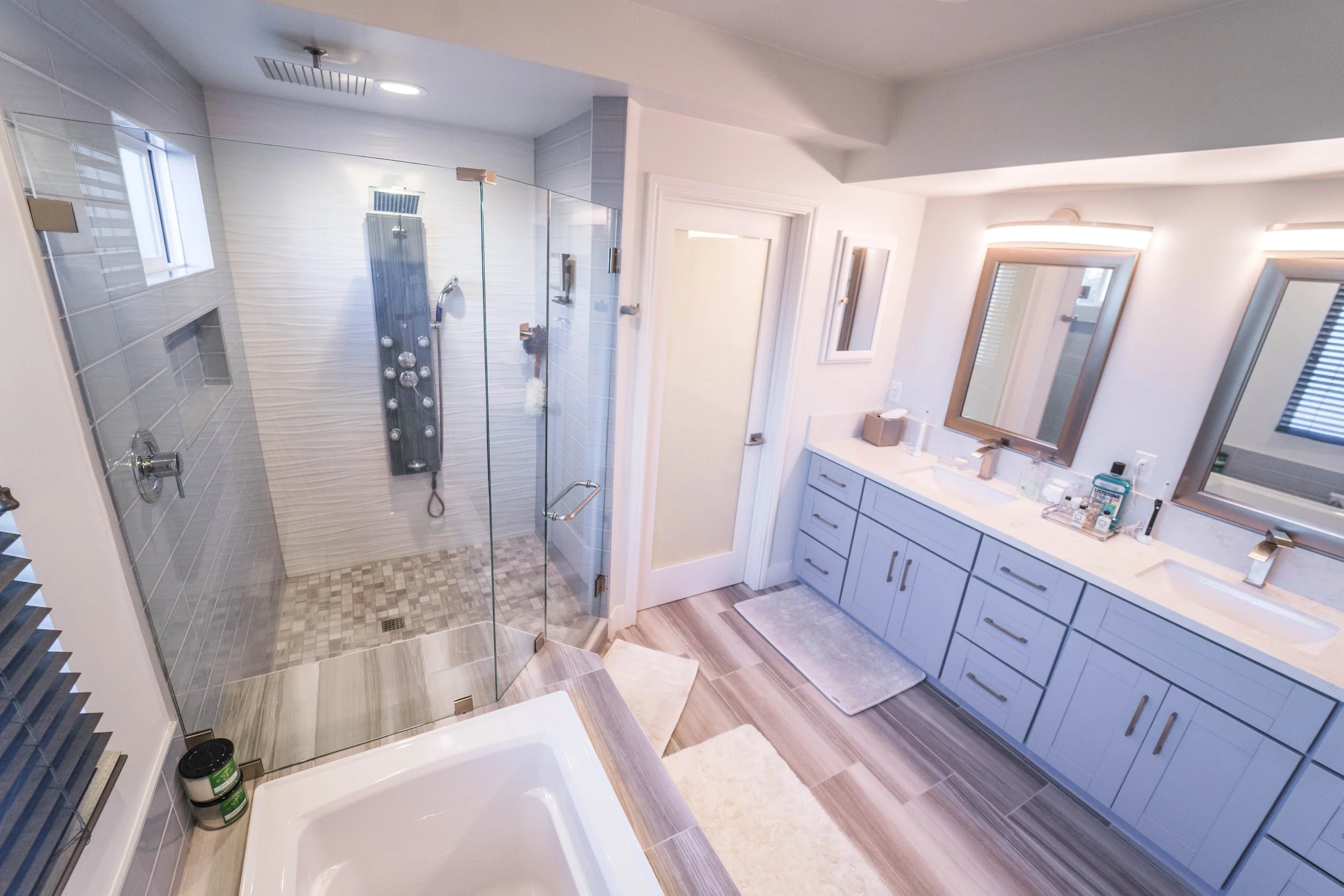 A modern bathroom equipped with a walk-in glass shower stall, a white vanity with a large mirror, and various toiletries on the countertop. The bathroom has wood-look tile flooring, white walls, and a window providing natural light.