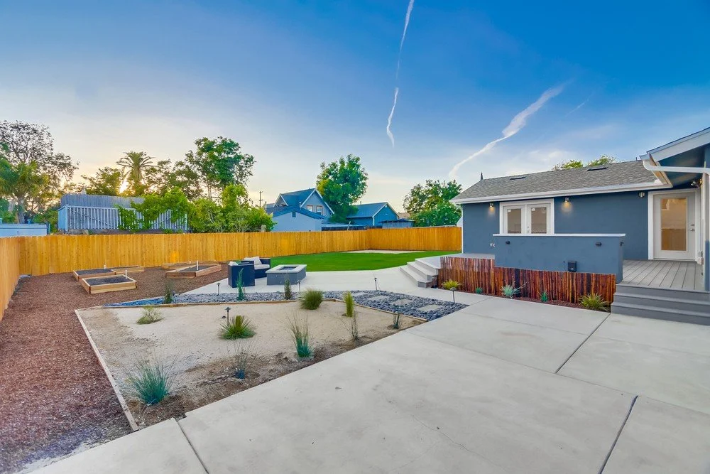 Backyard with a gray house, concrete patio, landscaped garden beds, fire pit, and a freshly mowed lawn, with a wooden fence and trees in the background under a blue sky.