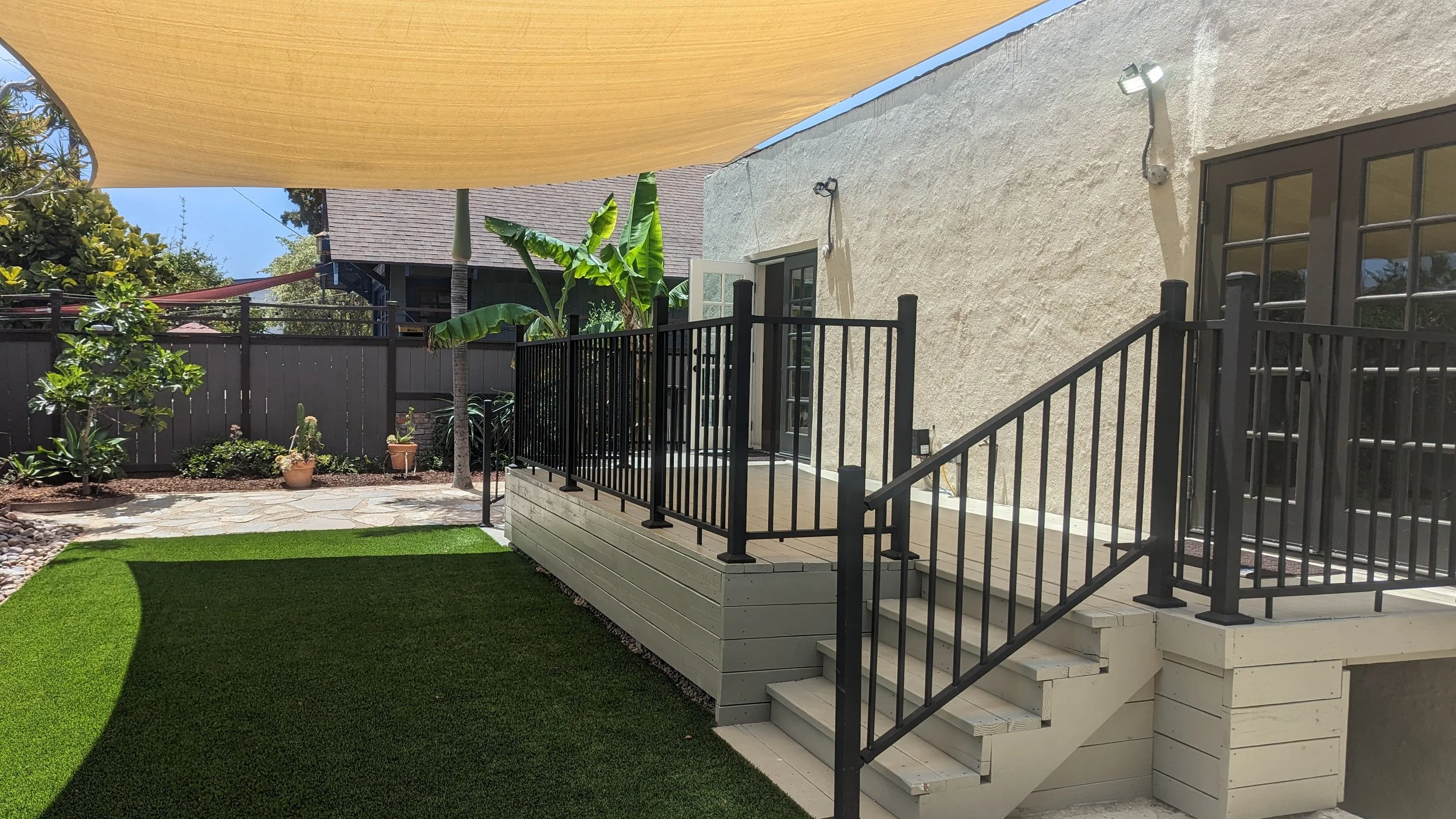 Backyard with a small wooden deck, black metal railing, steps, lush green grass, potted plants, banana tree, and a beige house wall with outdoor lights.
