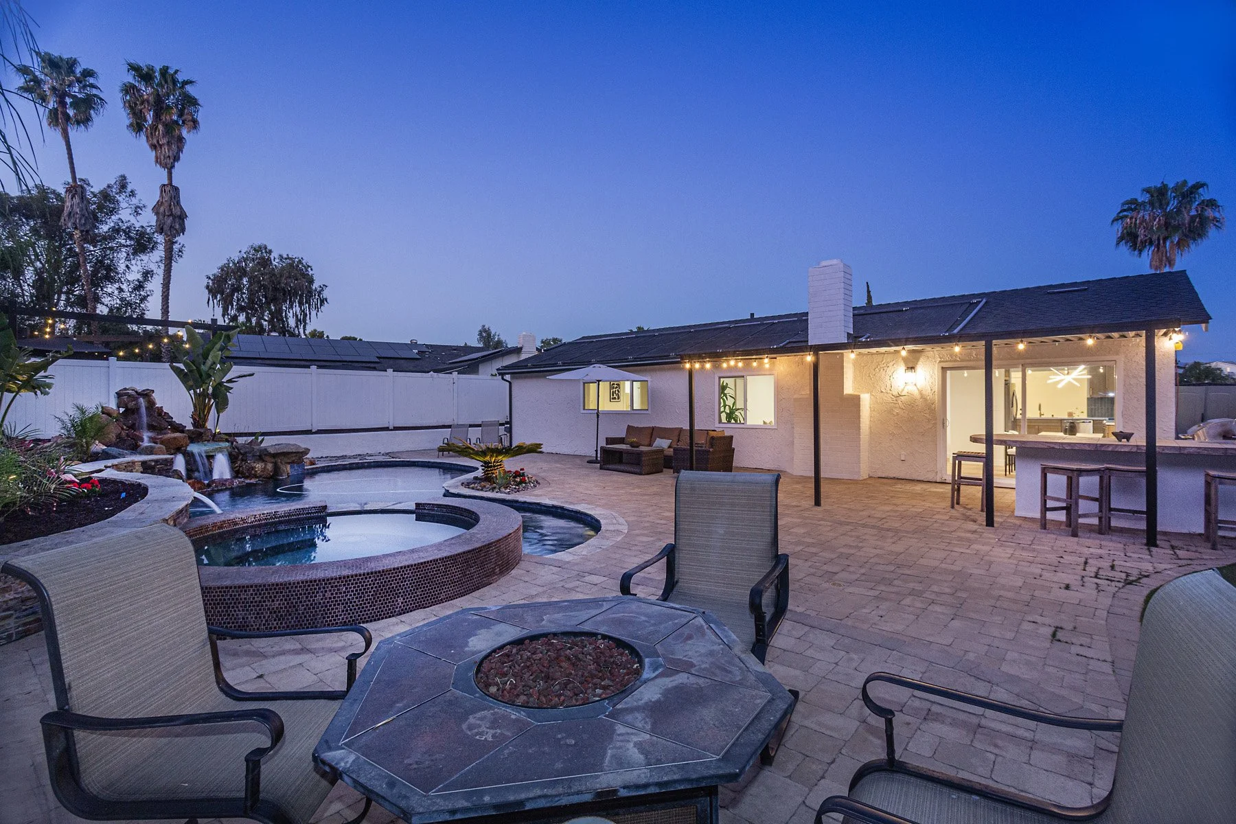 Backyard patio with a hot tub, fire pit, outdoor seating, water fountain, and string lights at dusk.