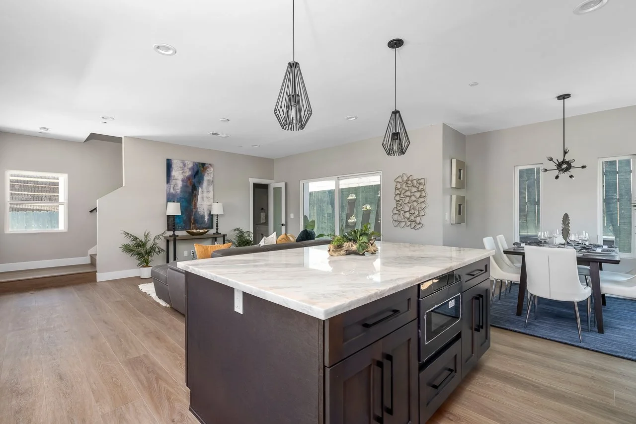 Modern open-concept living room and dining area with a kitchen island, white chairs, artwork, and large windows in a bright, neutral-colored home.