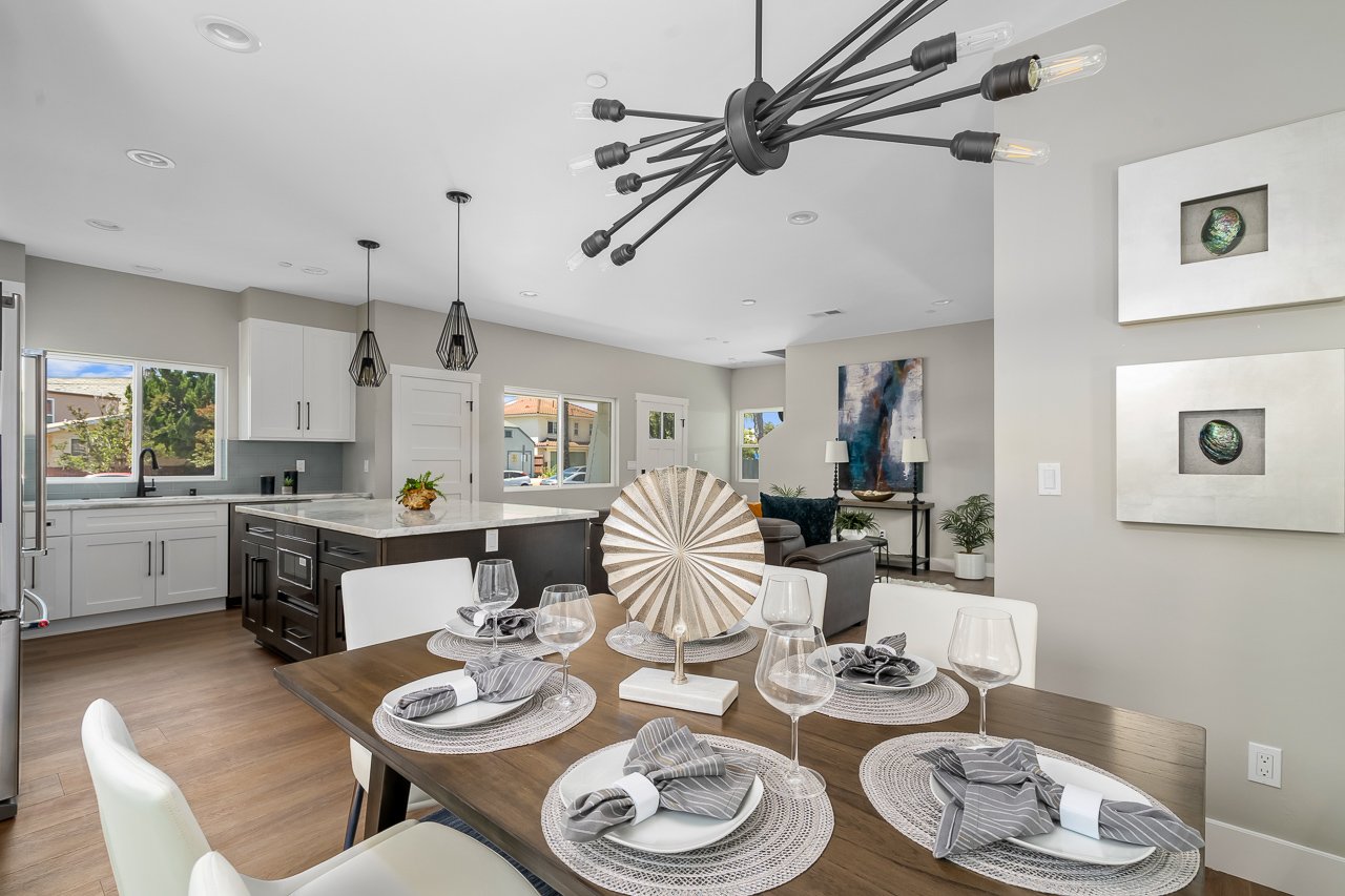 Modern open-concept dining and kitchen area with a wooden dining table set with wine glasses, napkins, and a decorative fan centerpiece, white cabinetry, black-and-white pendant lights above kitchen island, and contemporary wall art.