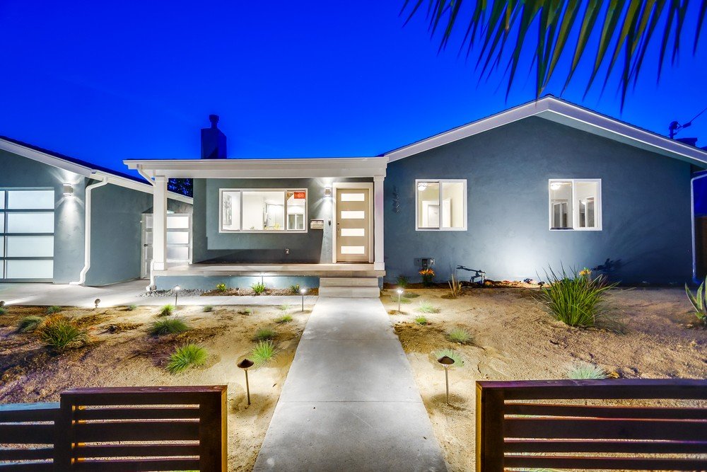 Modern single-story house at night with exterior lighting, gray exterior walls, a front door with horizontal windows, and a landscaped front yard with small plants and pathway lights.