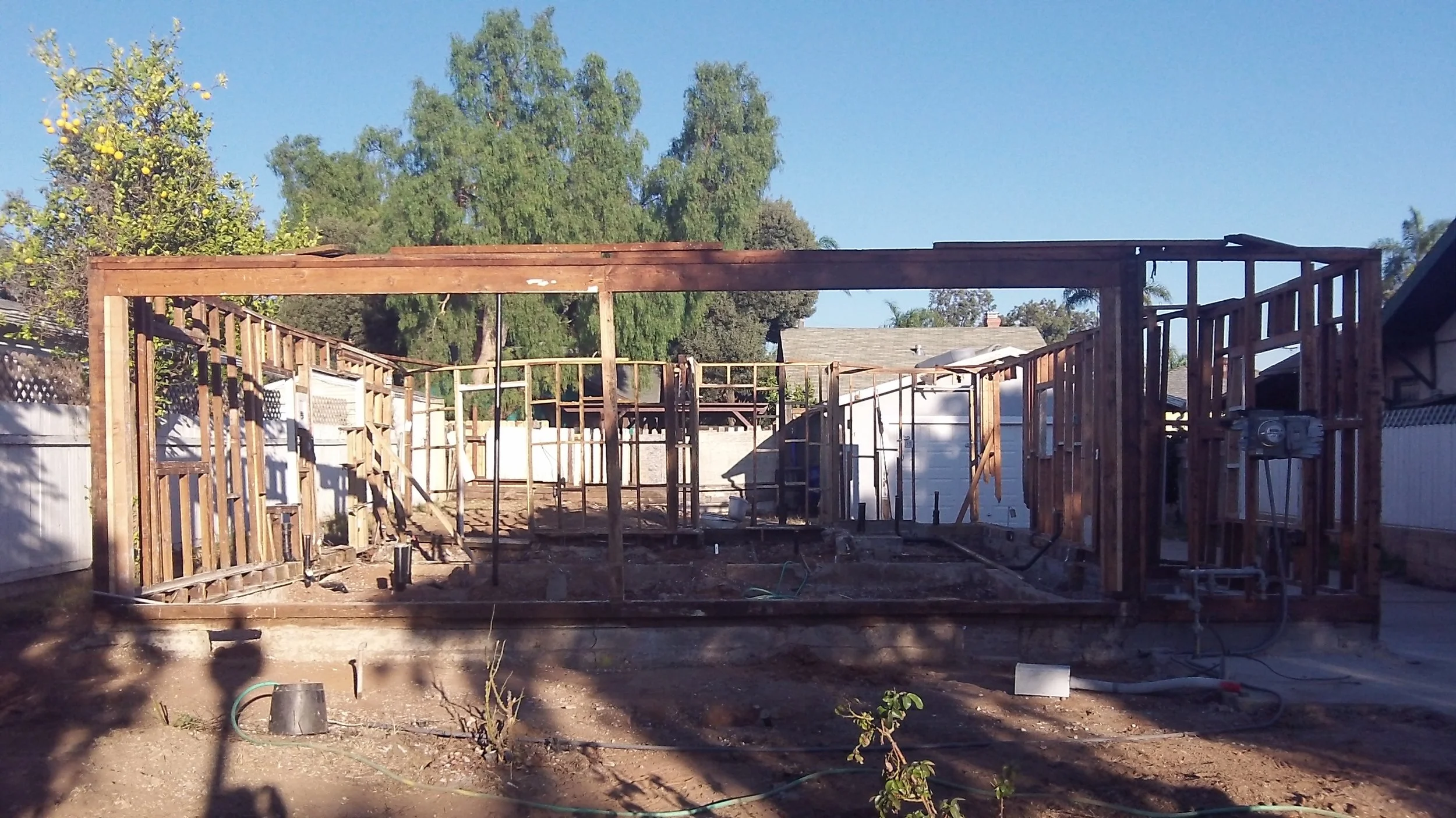 The image shows a backyard in the process of construction, with the wooden framing of a structure being built. The background includes trees and neighboring houses, with a clear blue sky overhead.