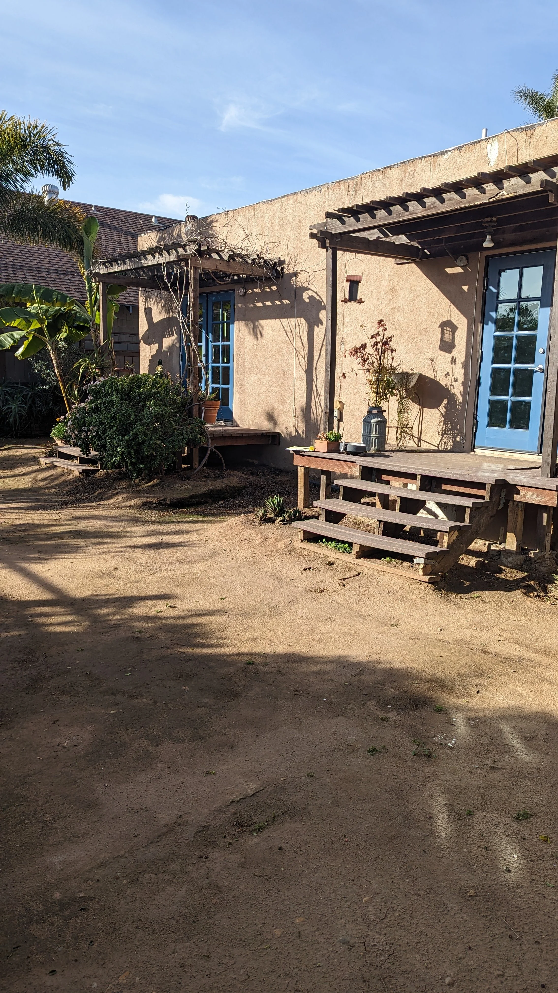 A backyard scene with a rustic house featuring a blue door, a wooden porch, and potted plants. There are trees and shrubs, with a dirt ground and a clear sky overhead.