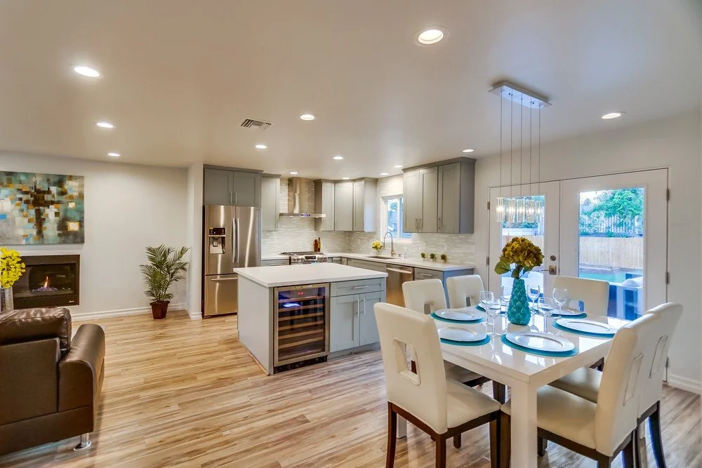 Open-concept kitchen and dining area with hardwood floors, gray cabinets, stainless steel appliances, and a glass dining table with six white chairs, decorated with a blue vase and yellow flowers.