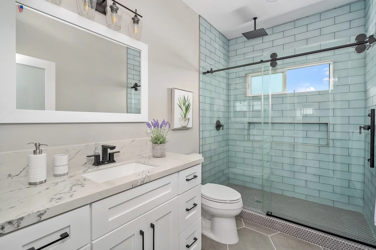 Modern bathroom with a white vanity, marble countertop, black fixtures, a potted lavender plant, a framed botanical print, a toilet, and a walk-in shower with light blue subway tiles and a window showing a sunny sky.