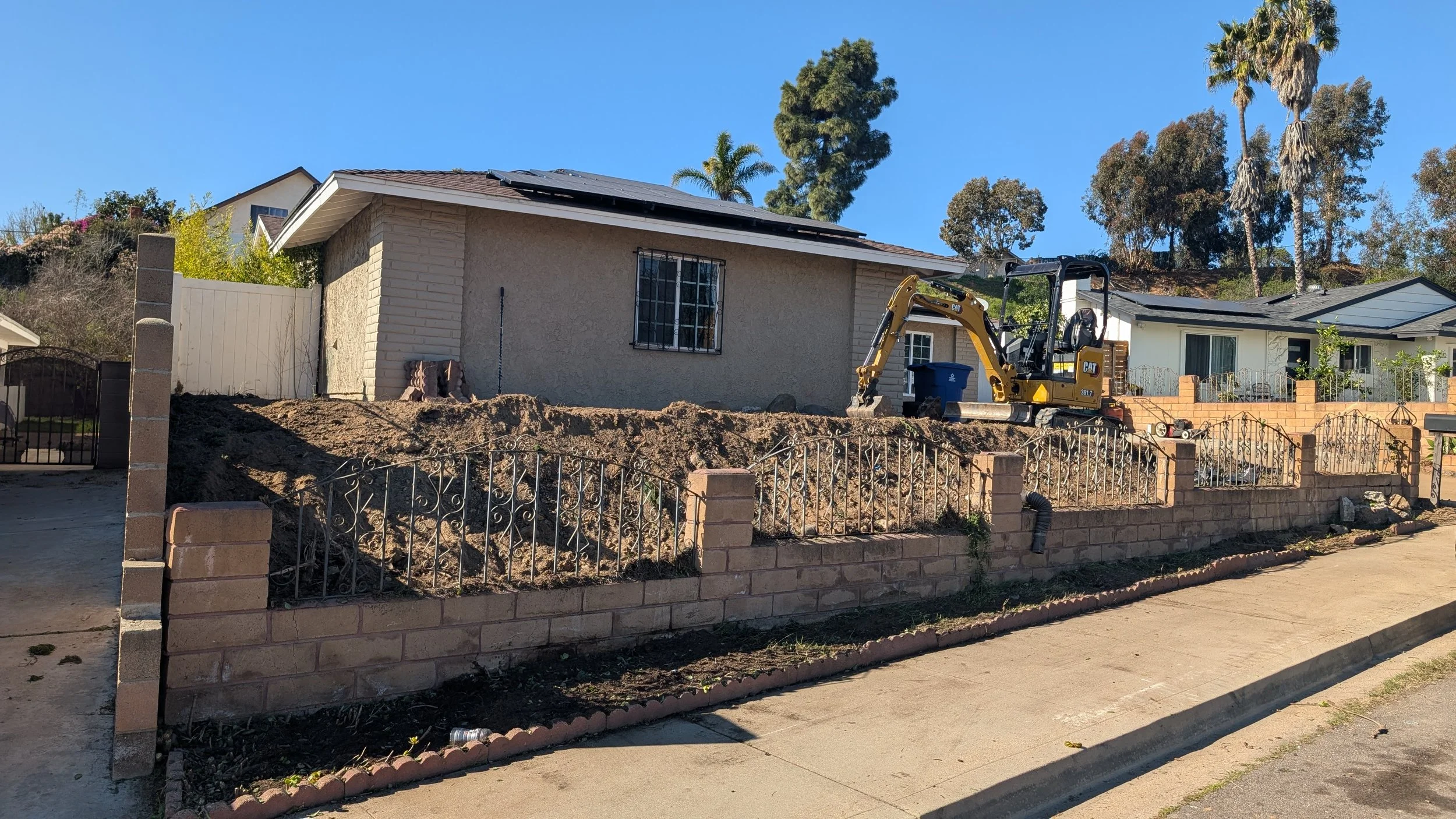 Residential backyard under renovation with a small excavator, dirt pile, and a brick and metal fence, with houses and tall trees in the background.
