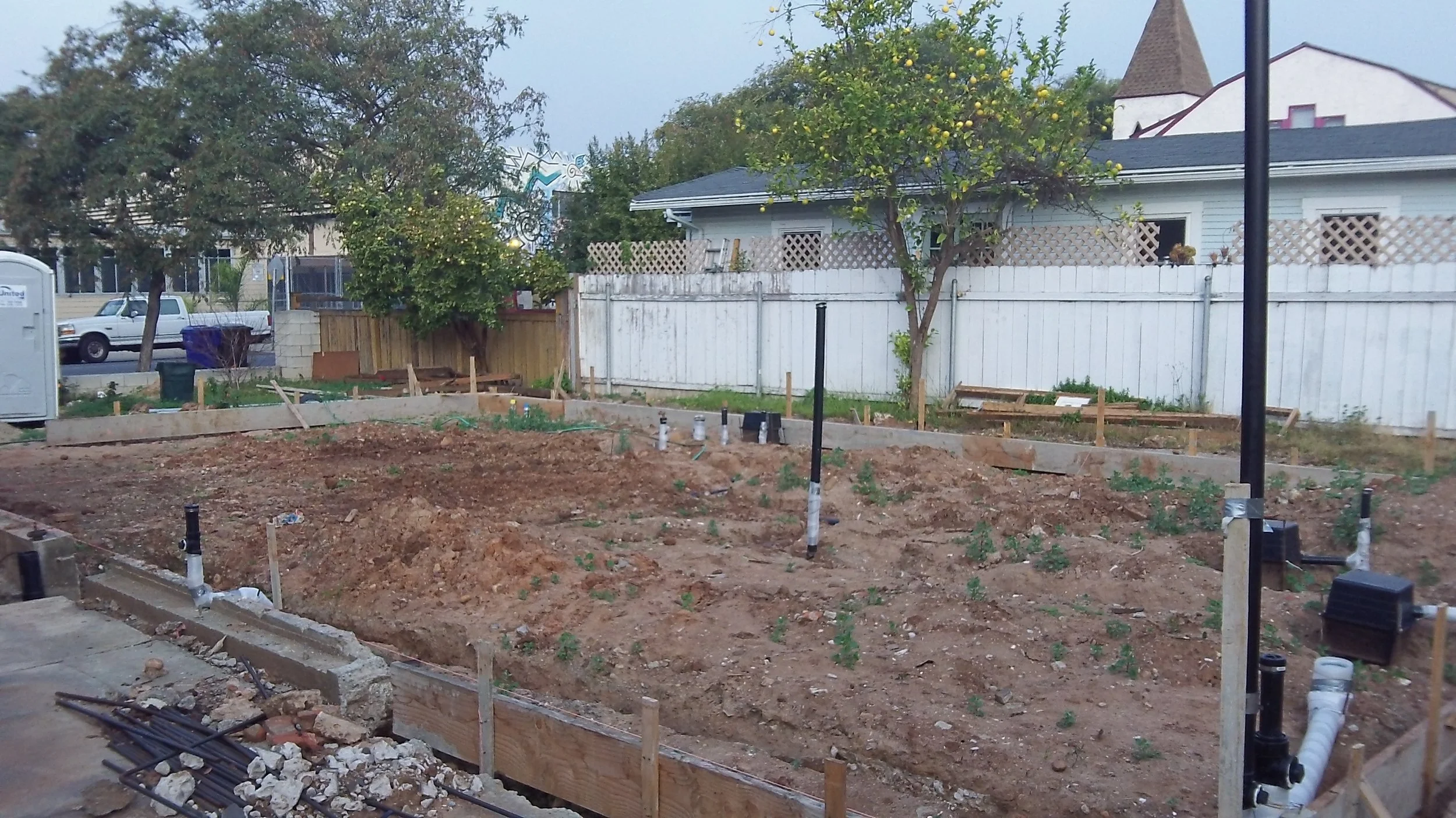 A backyard garden starting to be landscaped with wooden borders, pipes for irrigation, and bare earth. There are trees and fences, along with houses and a street visible in the background.
