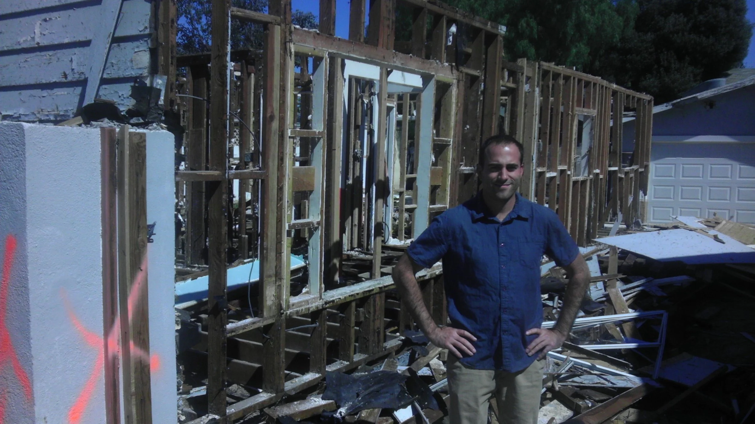 A man stands with his hands on his hips in front of a house under construction, with exposed wooden framing and some debris around.