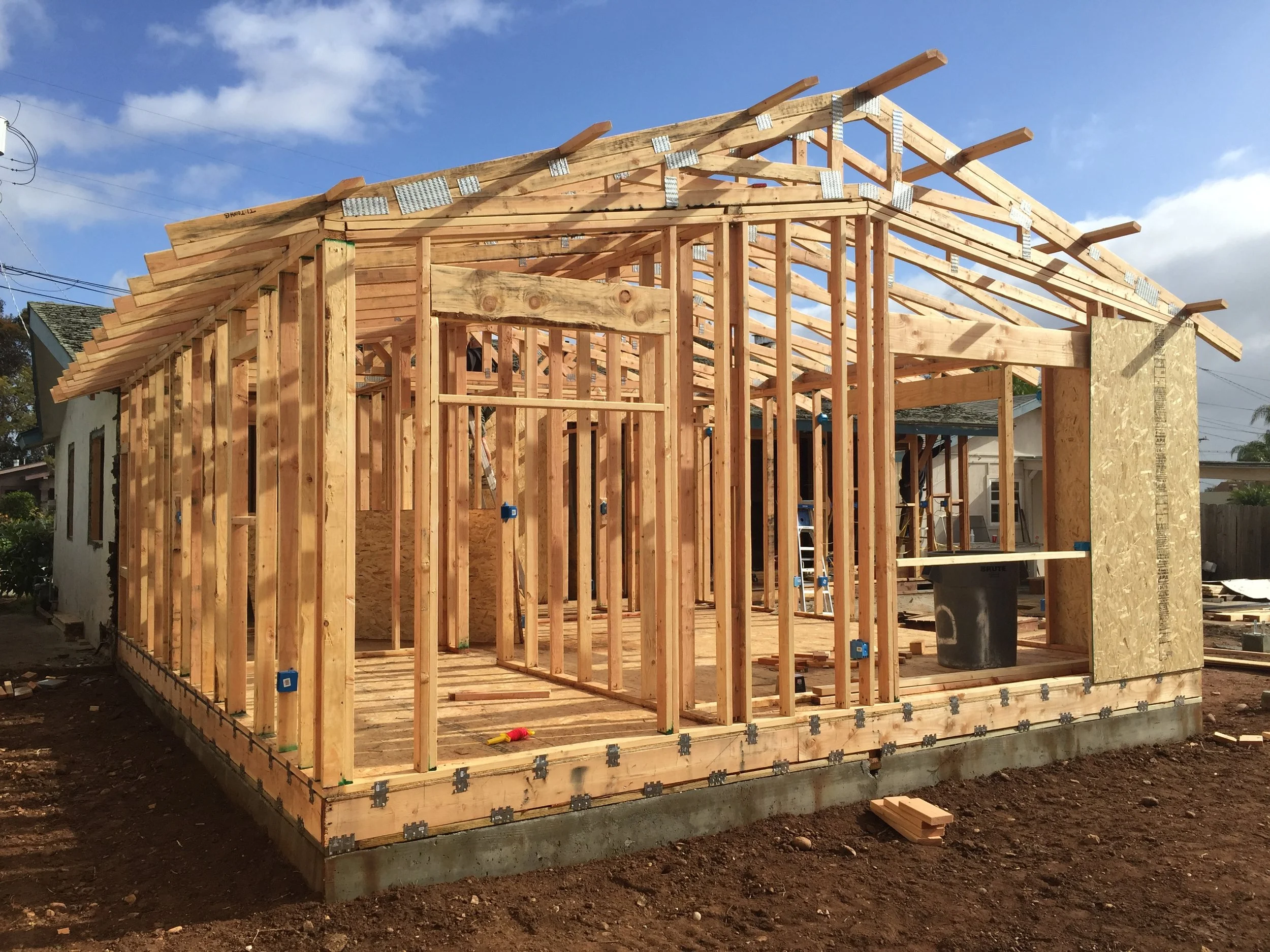 Wooden house framing under construction with a partly built roof structure, against a background of blue sky with clouds.