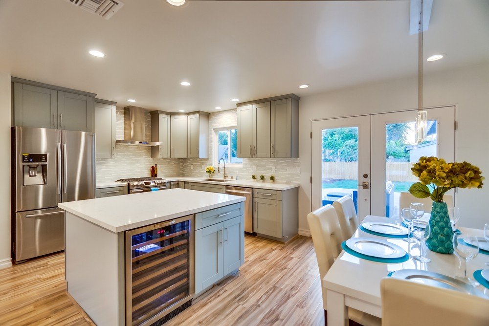 Modern kitchen with light gray cabinets, white countertops, stainless steel refrigerator, and a dining area with a table set for four and a vase of flowers.