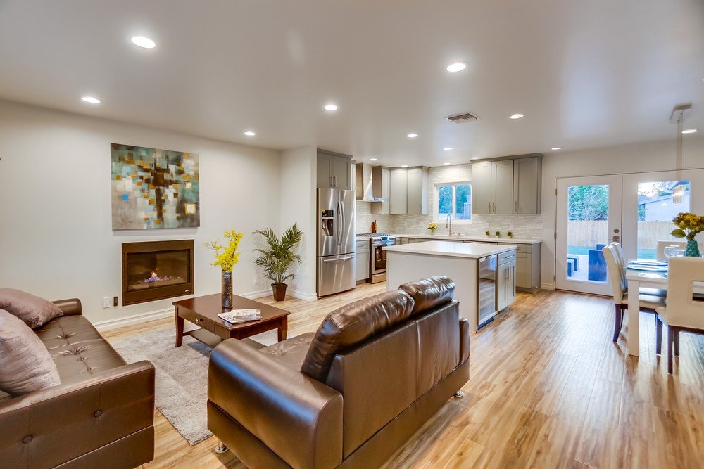 Open-concept living room and kitchen with leather sofas, fireplace, wooden flooring, kitchen island, stainless steel appliances, and dining table with chairs, near sliding glass doors.
