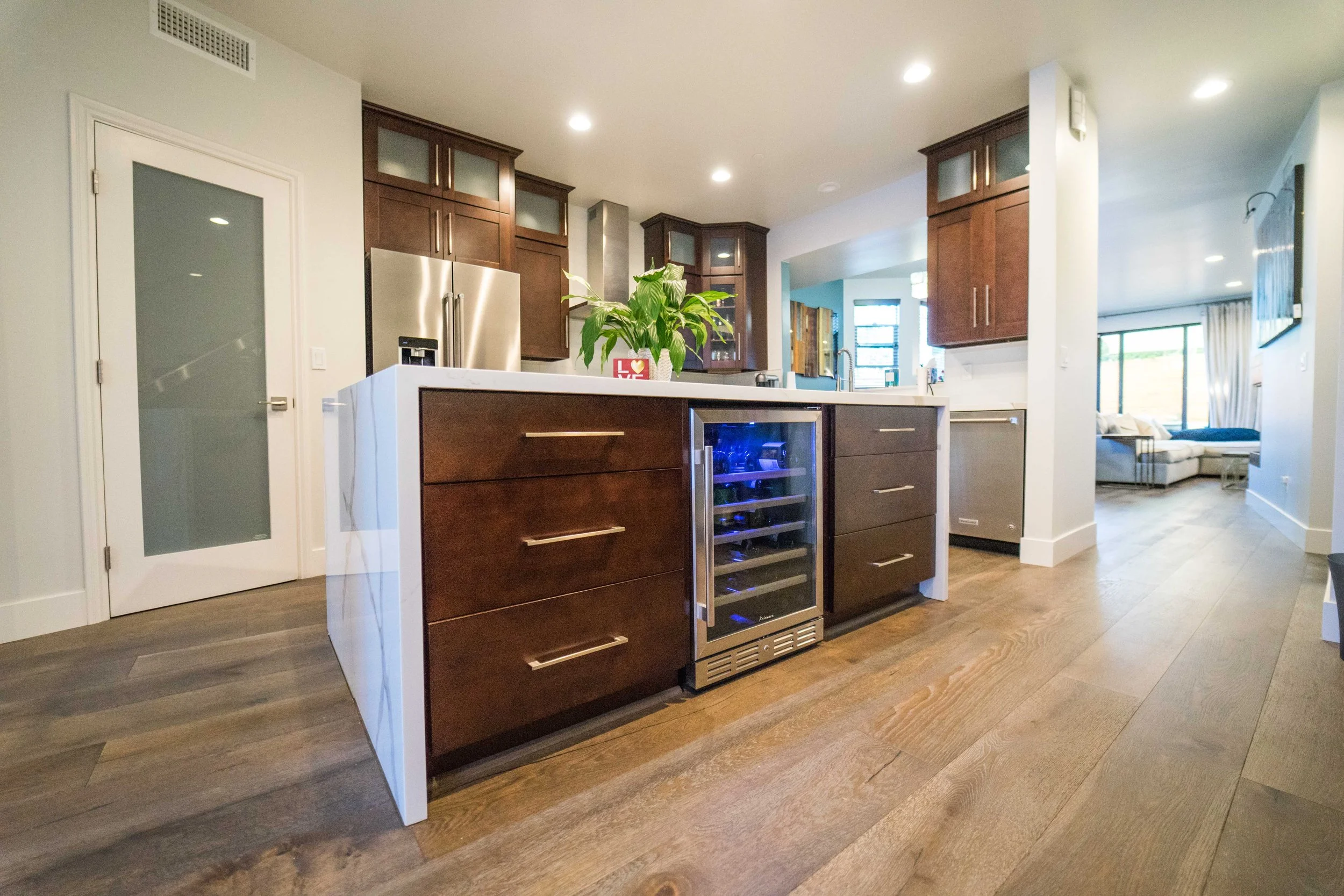 Modern kitchen with dark wood cabinets, white countertops, stainless steel refrigerator, and built-in wine cooler. A green plant and a red decorative box are on the counter. Bright lighting and hardwood floors are visible.