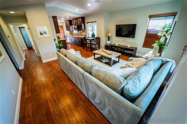Living room with beige sectional sofa, wooden floors, wall-mounted TV, windows with blinds, and a nearby kitchen area.