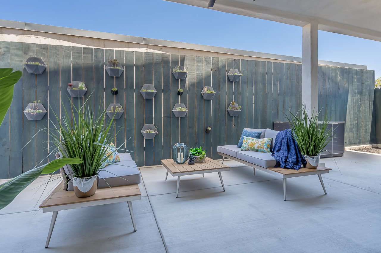 Modern outdoor patio with two cushioned lounge chairs, a wooden coffee table, large potted plants, and a decorative wall with hanging planters, under a roof extension, against a blue sky.