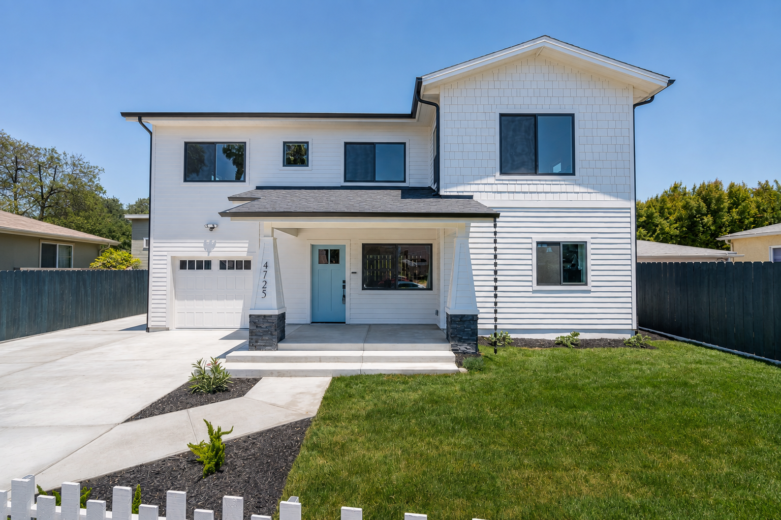 A modern two-story house with white siding, black window frames, and a light blue front door. There is a garage on the left, a small porch with stairs, a well-maintained lawn, and a white picket fence in the front yard. The sky is clear and blue.