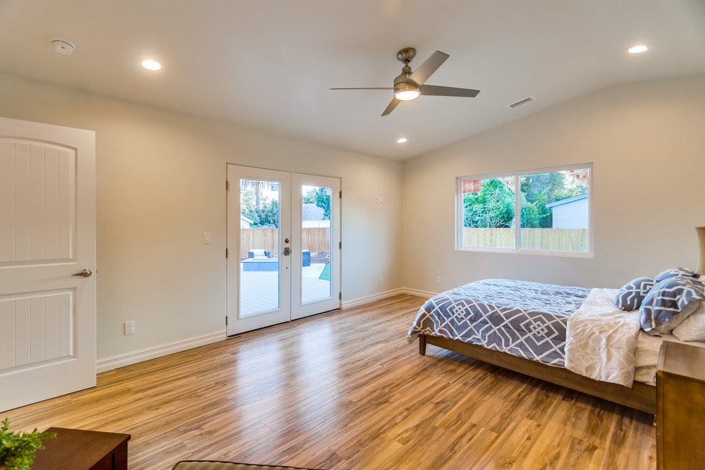 A bedroom with a wooden floor, a bed with blue and white bedding, a window showing outdoor trees, and French doors leading outside.
