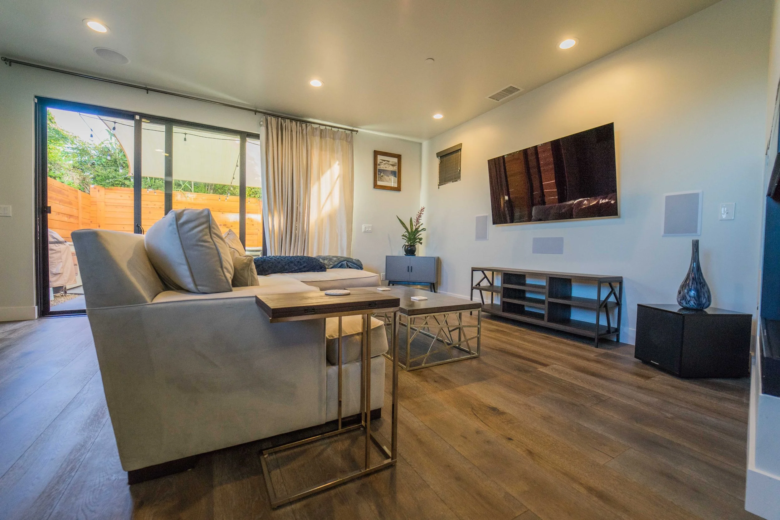 Living room with white sofa, coffee table, flat-screen TV on the wall, sliding glass door with curtains, wooden floor, and decorative vase.