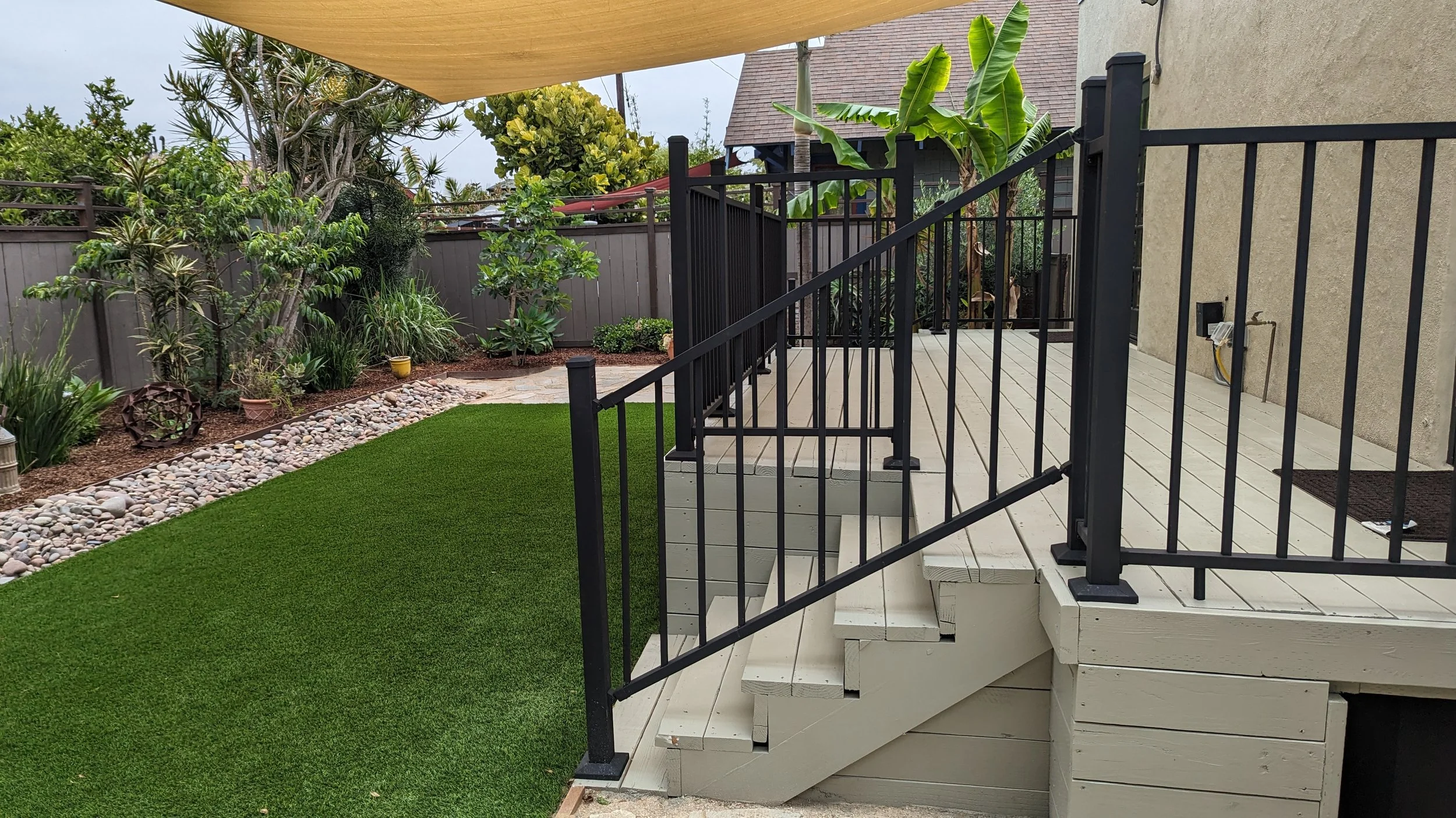Backyard patio with stairs, black metal railing, wooden deck, green artificial turf, landscaped garden with trees, shrubs, rocks, and potted plants, beige wall, and house roof visible in the background.