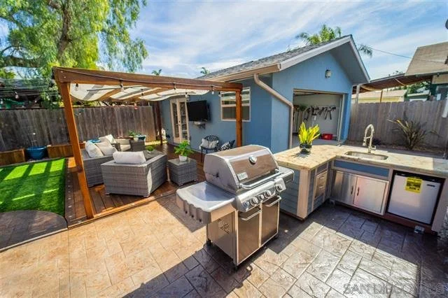 Backyard patio with outdoor kitchen and seating area, including a grill, a countertop with a sink, a house with a blue exterior, a shaded lounge area with furniture, and a green lawn with trees.