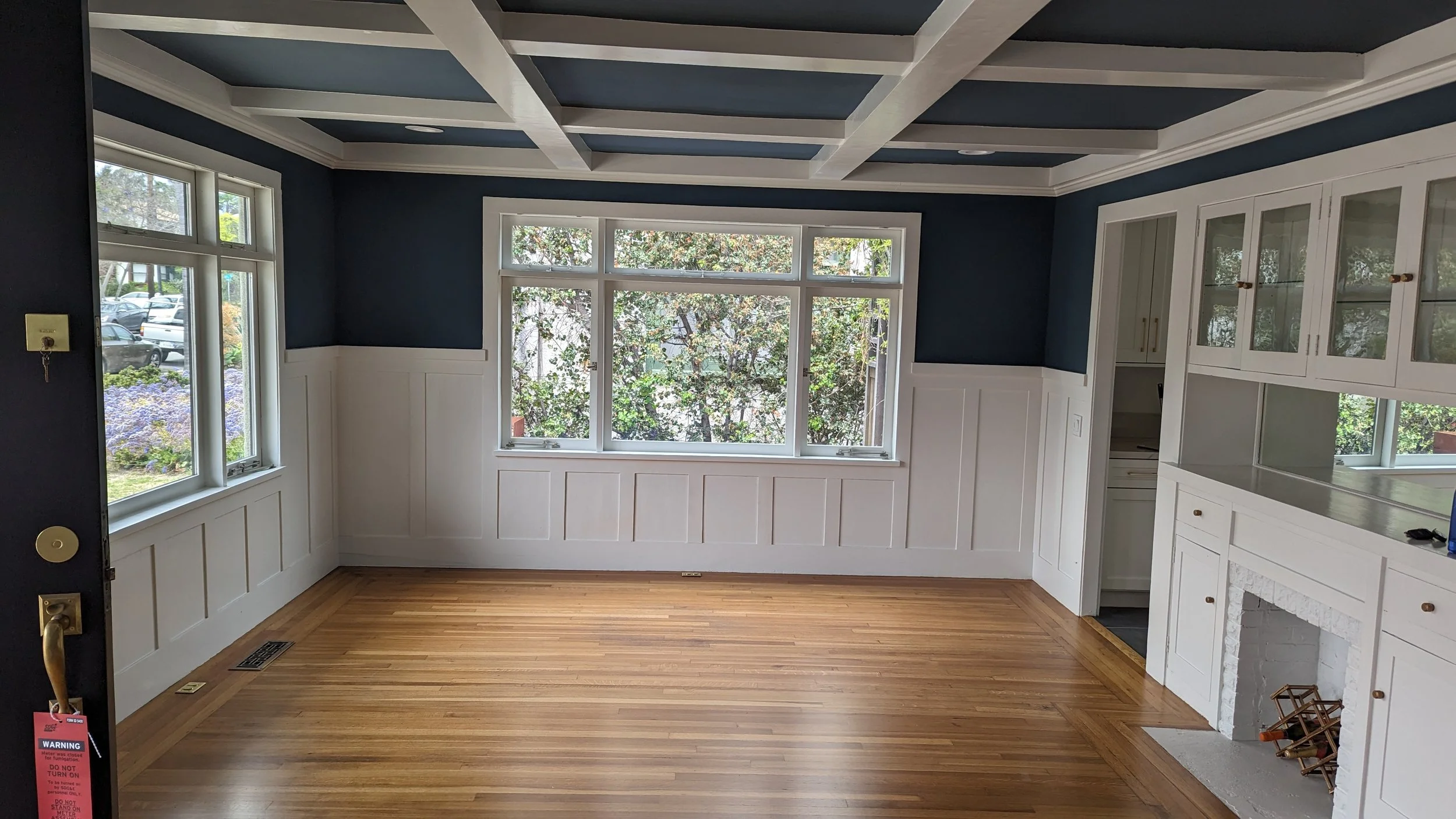 Empty room with large windows, dark blue walls, white wainscot paneling, built-in white cabinets with glass doors, hardwood floor, and a white fireplace.
