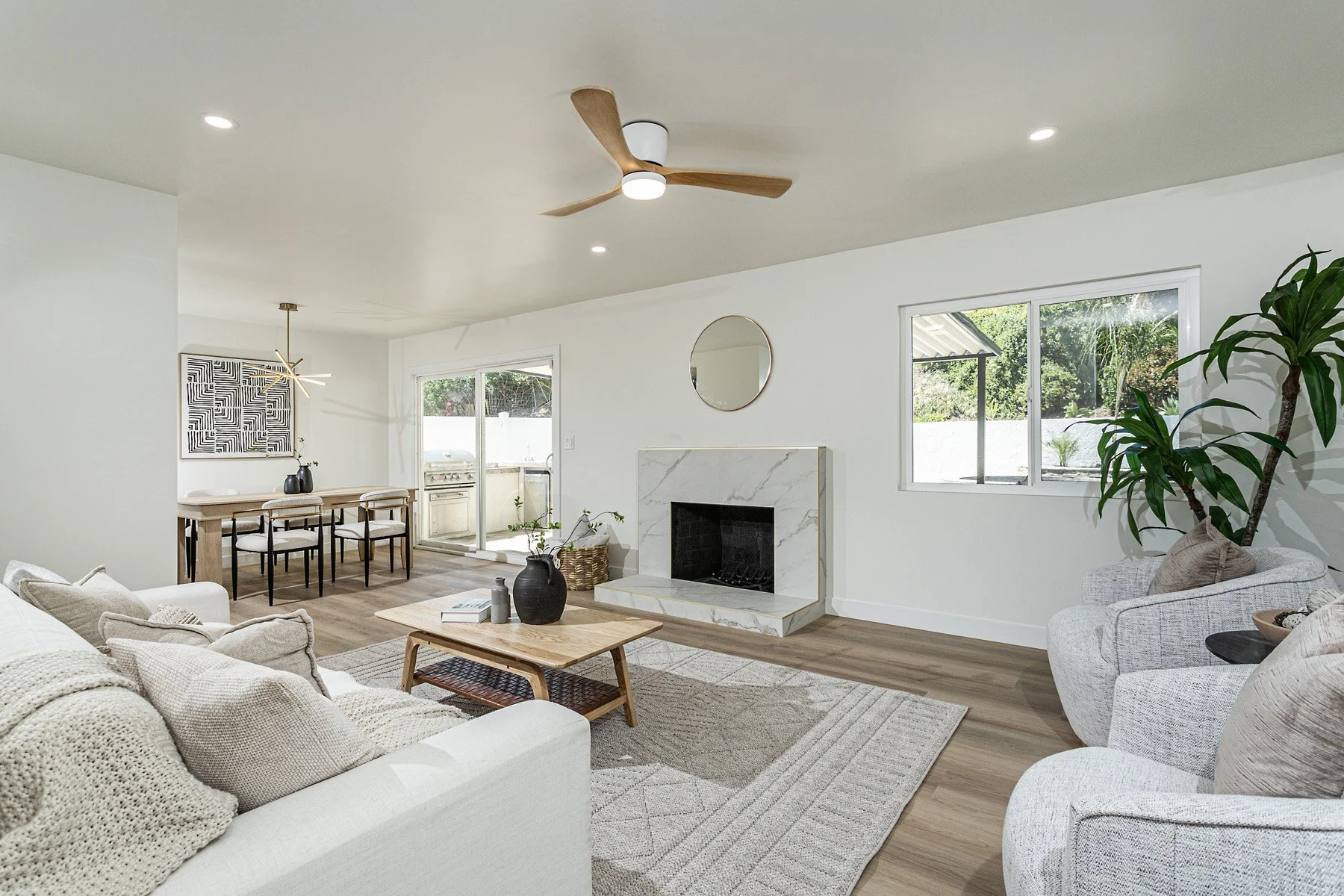 Open-concept living room with white walls, beige sofas, a wooden coffee table, and a fireplace with a white marble mantle. There are large windows, a potted plant, and a dining area with a wooden table and black chairs in the background. A ceiling fan with wooden blades is installed on the ceiling.