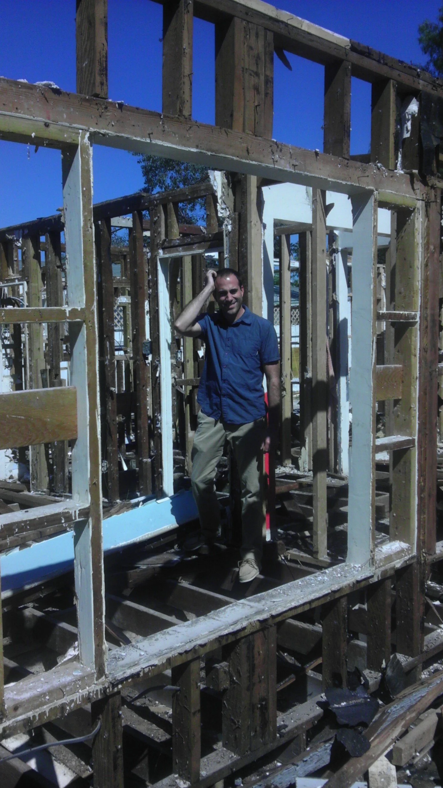 A man standing inside the wooden frame of a house under construction, smiling and touching his head, with a bright blue sky in the background.
