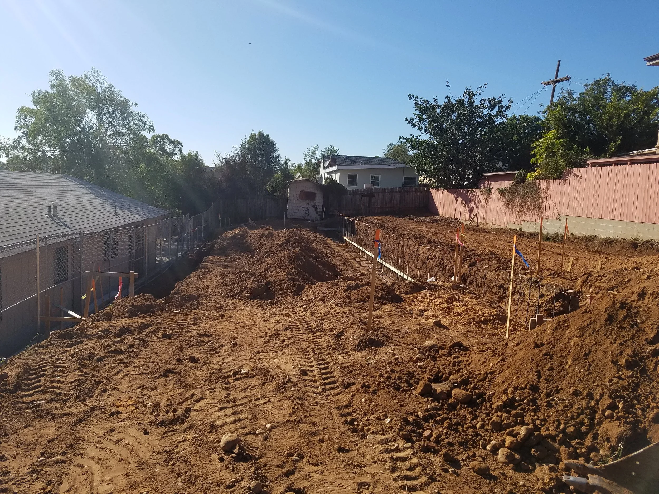 A construction site with excavated ground, wooden stakes, and construction fencing, with trees, houses, and a clear blue sky in the background.