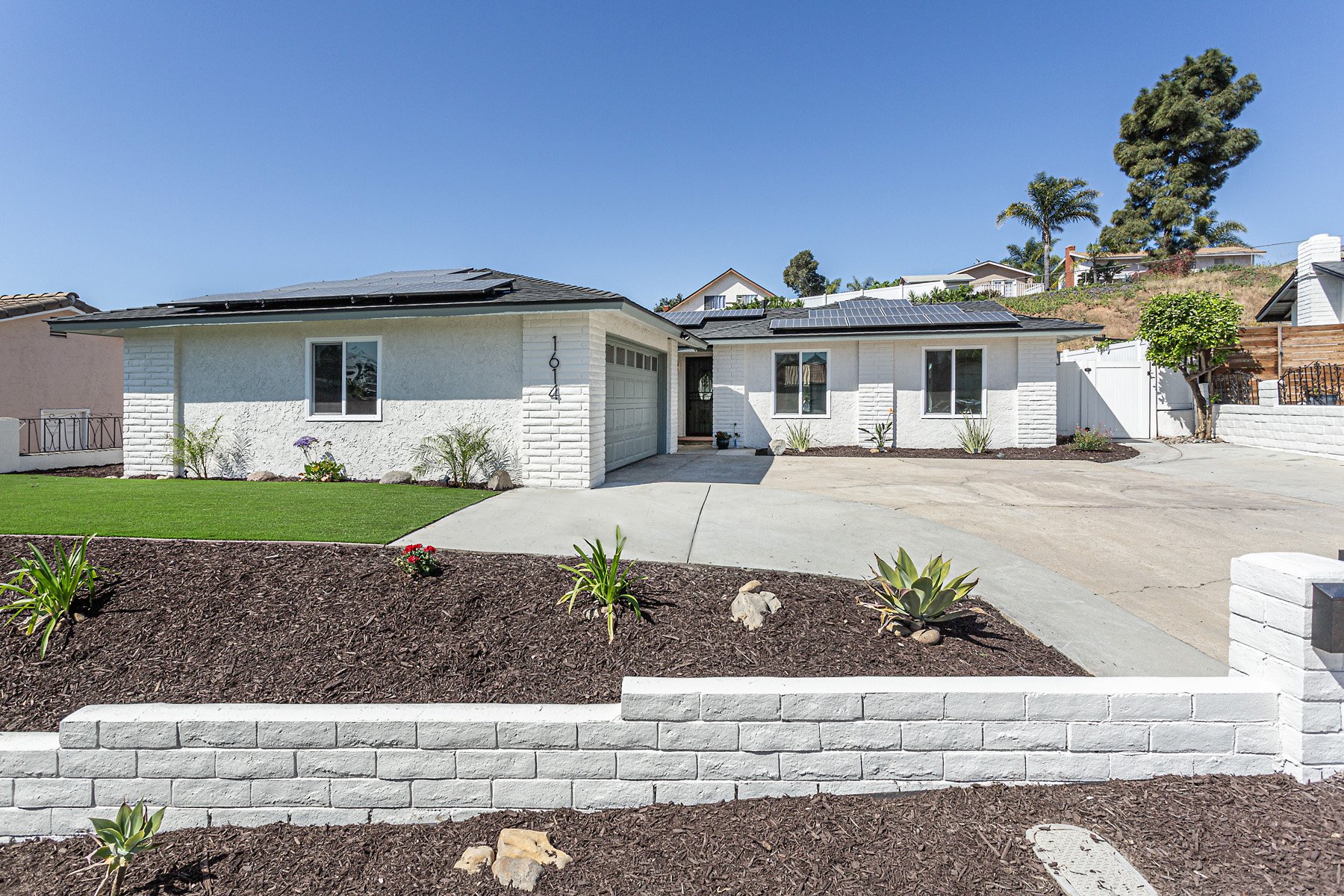 White house with solar panels on the roof, a gray driveway, landscaped yard with green grass, small plants, and mulch, surrounded by a white brick wall, under a clear blue sky.