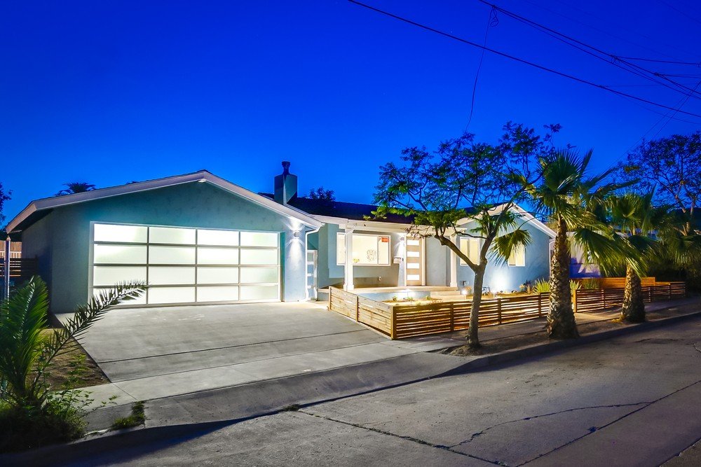 Modern house with lit garage, wooden fencing, and palm trees in a residential neighborhood at dusk.