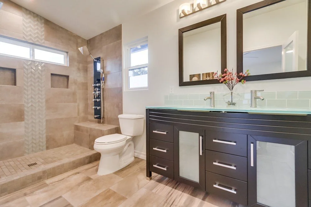 Modern bathroom with beige tile walk-in shower, white toilet, black vanity with two sinks, large mirrors, and pink flowers in a vase.