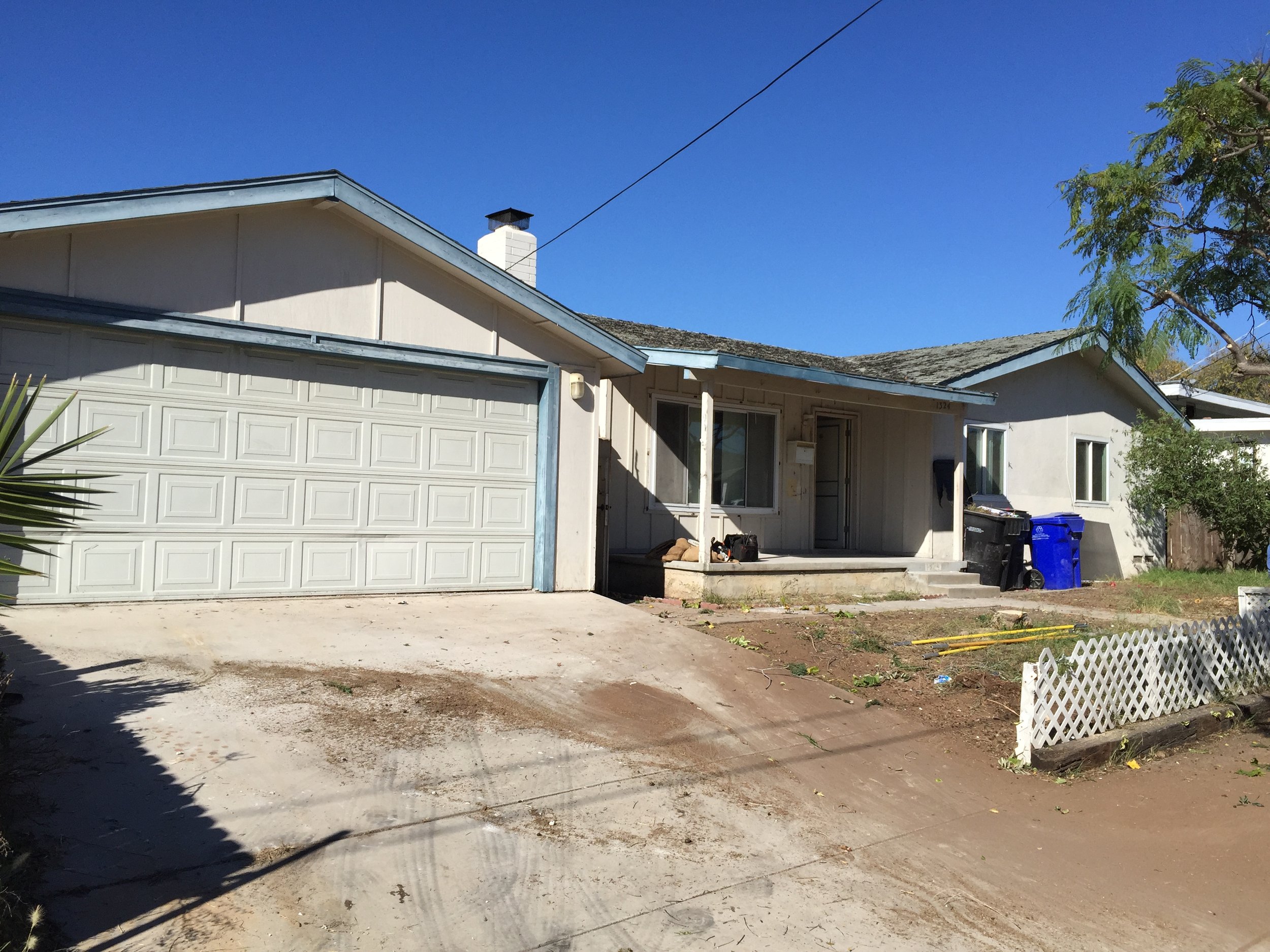Front of a beige house with a two-car garage, some turf and dirt in front, and a small porch with items on it. A blue recycling bin and a black trash bin are beside the house. A small white fence is in the yard, and trees and a clear blue sky are in the background.