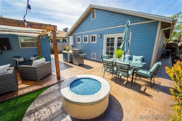 Backyard patio with outdoor seating, a hot tub, and a dining table with chairs next to a blue house with a sloped roof and multiple windows.