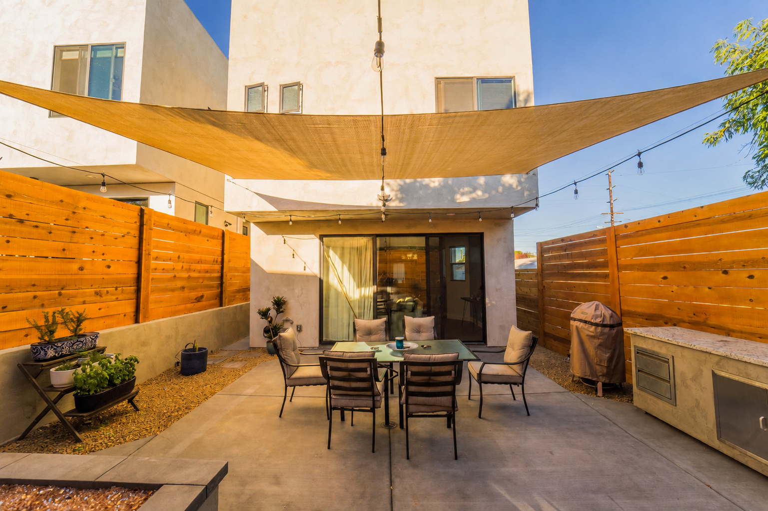 Backyard patio with outdoor dining table, six chairs, potted plants, wooden fencing, and a beige sunshade sail